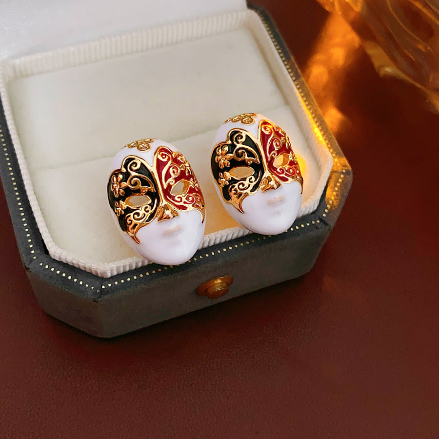 Enamel masquerade face stud earrings with red and black halves and gold filigree on a velvet jewelry box — Venetian Masquerade Studs.