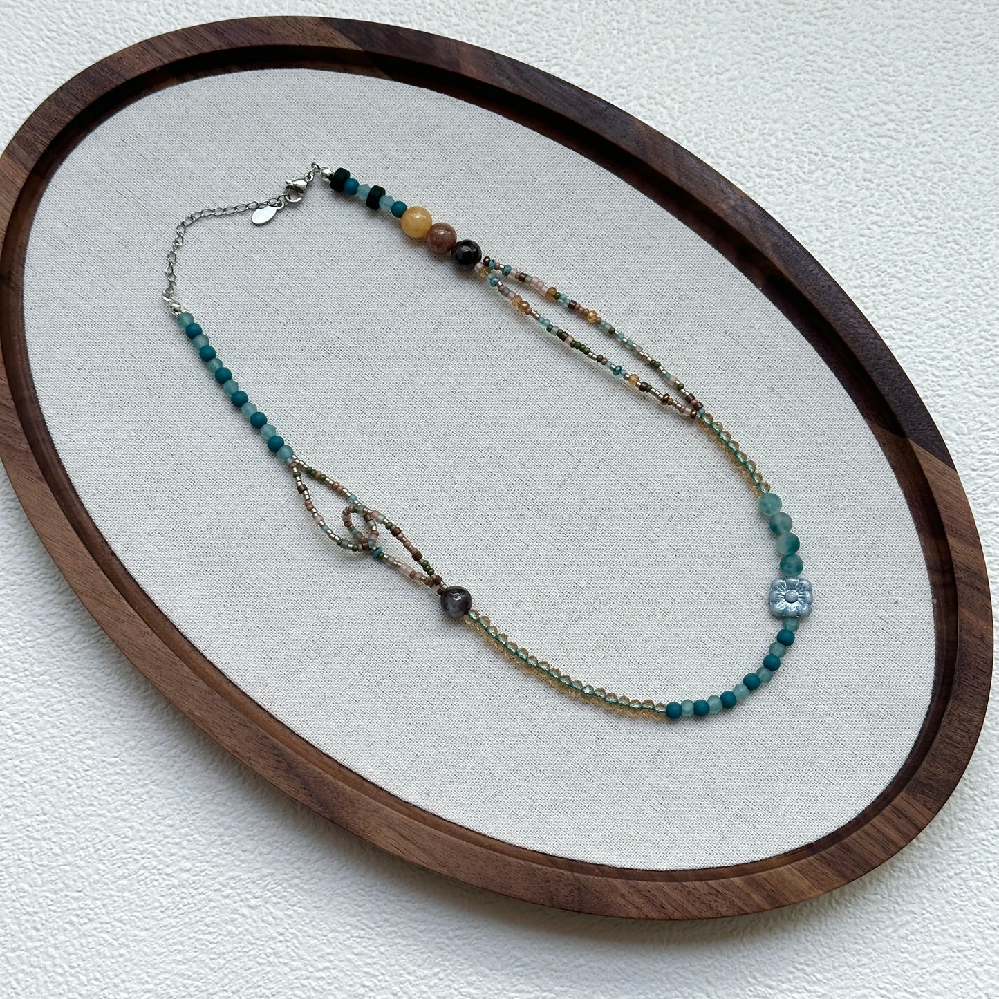 Aqua Loop Blossom Necklace displayed on an oval tray – serene lagoon blues with sandy-gold shimmer for a modern coastal look
