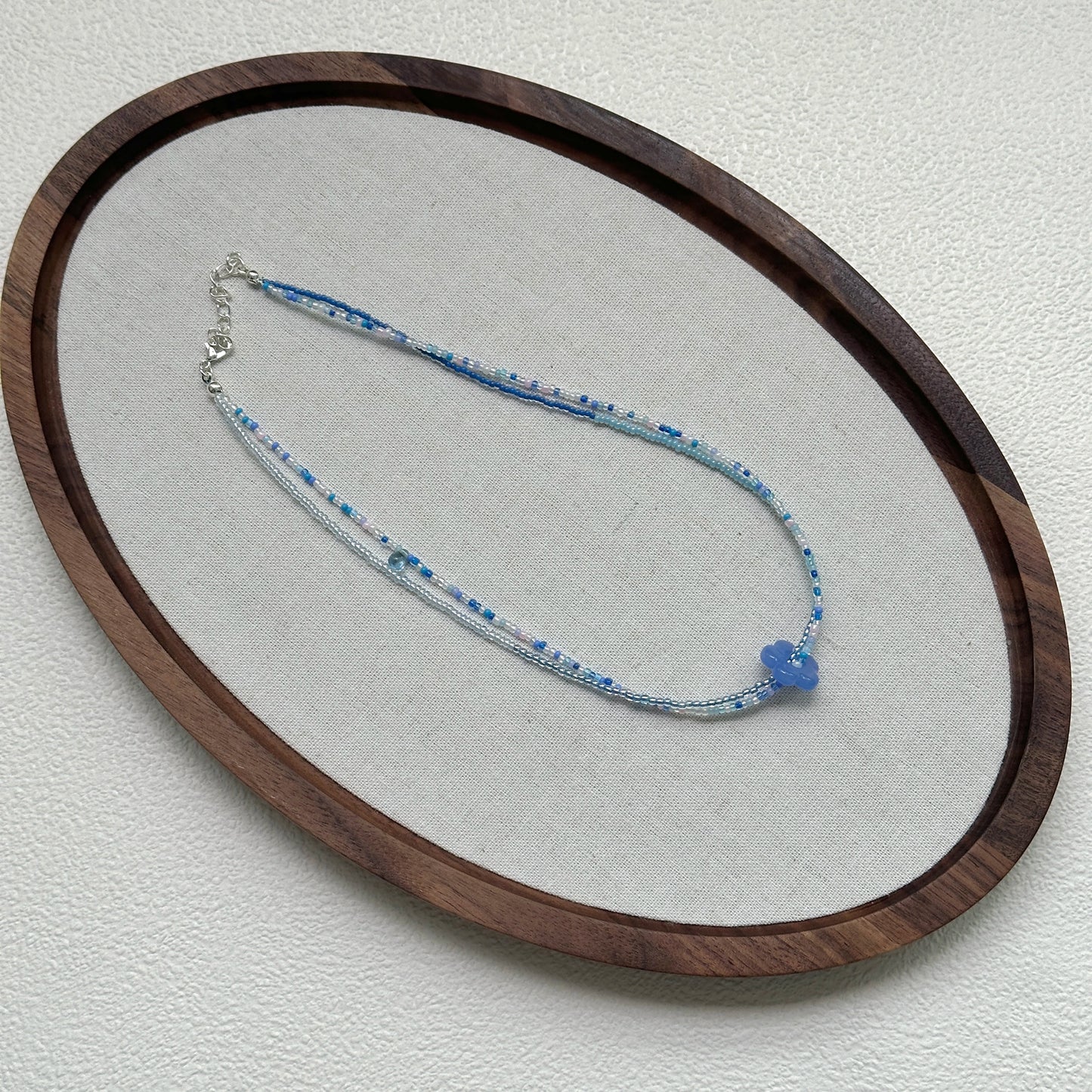 Azure Bloom Necklace oval flat lay – handcrafted minimal beaded necklace with azure flower charm and mixed blue-white beads by Glazenya