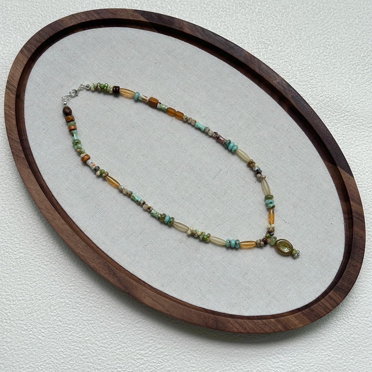 Verdant Amulet Necklace oval flat lay – handcrafted beaded necklace with amber-gold, green, and ivory beads surrounding a vintage pendant by Glazenya