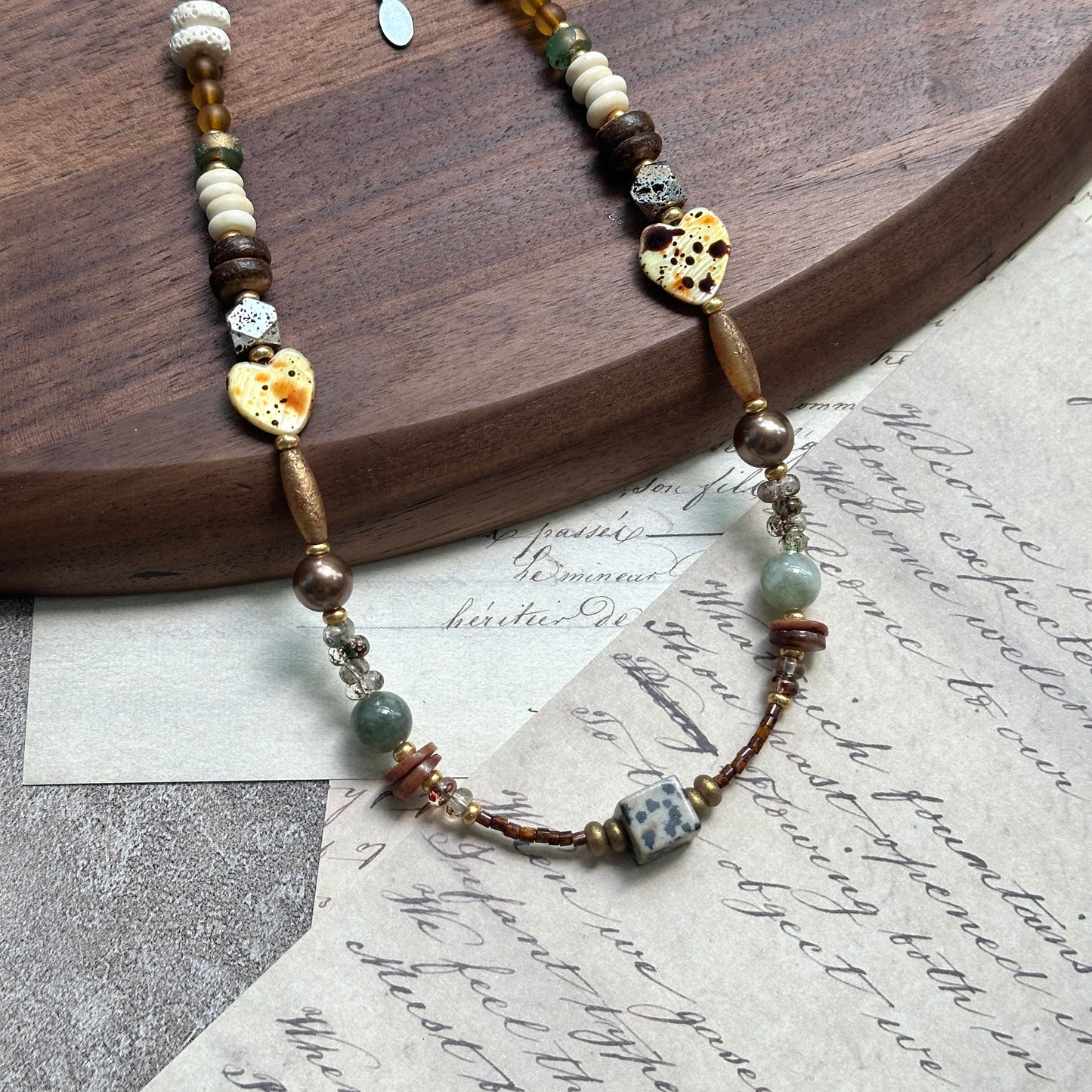 Sage & Saffron Necklace — angled view beside a wooden tray; close-up of speckled heart beads, amber tones, and dotted cube focal
