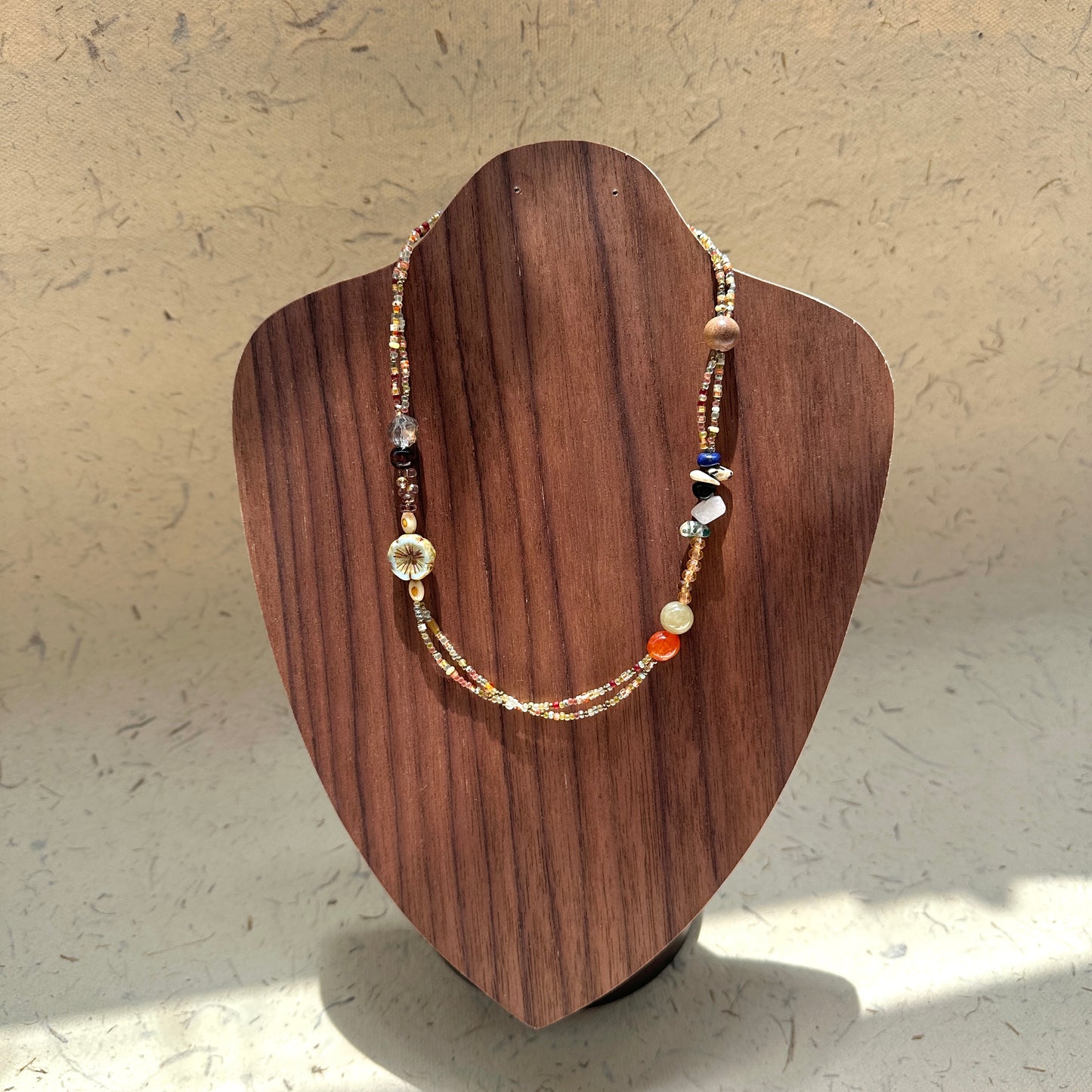 Marigold Mosaic Necklace on wood display bust — warm marigold and honey-amber beads with a floral coin and mixed accents in an asymmetric design