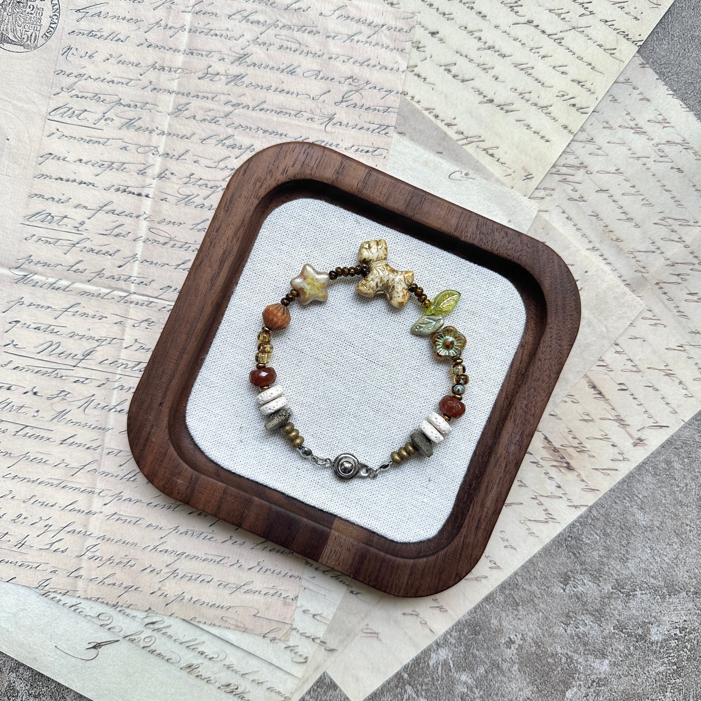 Meadow Pup Bracelet — flat lay in a wooden tray on vintage papers; warm amber and neutral beads with puppy, leaf, star, and floral accents