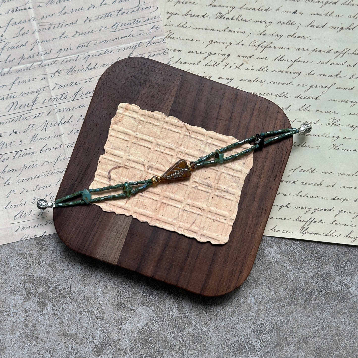 Verdant Talisman Bracelet — flat lay on textured papers; deep forest-green beads with a botanical heart talisman and magnetic clasp 4