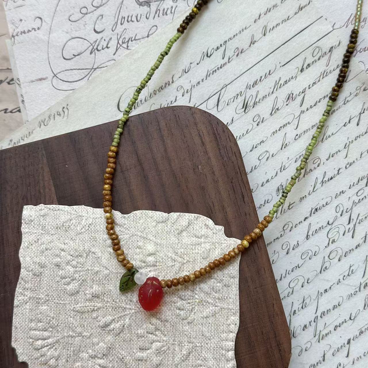 Wild Strawberry Necklace close-up on rustic paper, featuring glass strawberry pendant with petite green leaf, dewdrop accent, and moss-green beads