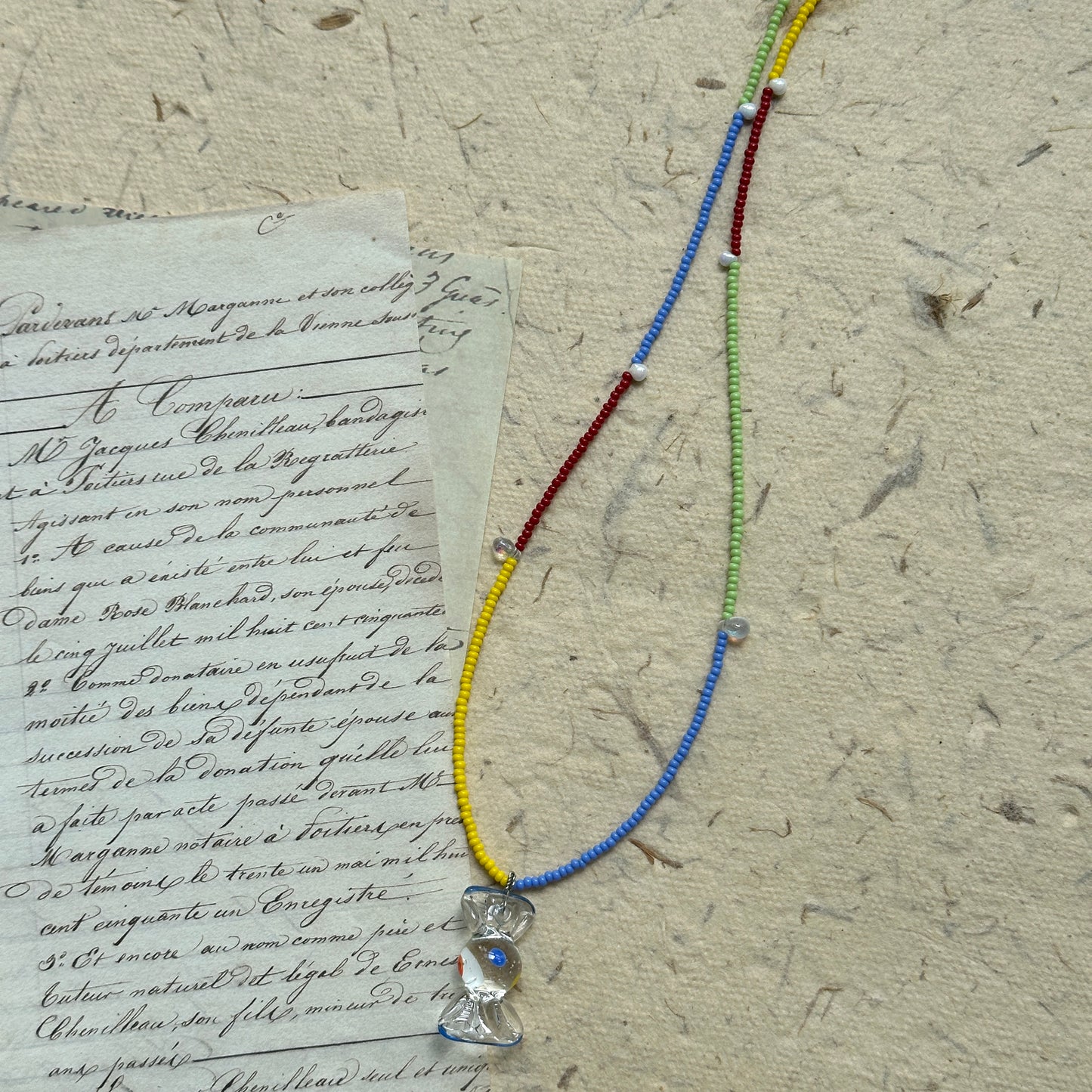 Handcrafted Joyful Hour Necklace with rainbow-colored seed beads and a crystal-like hourglass pendant, displayed flat on vintage paper background.