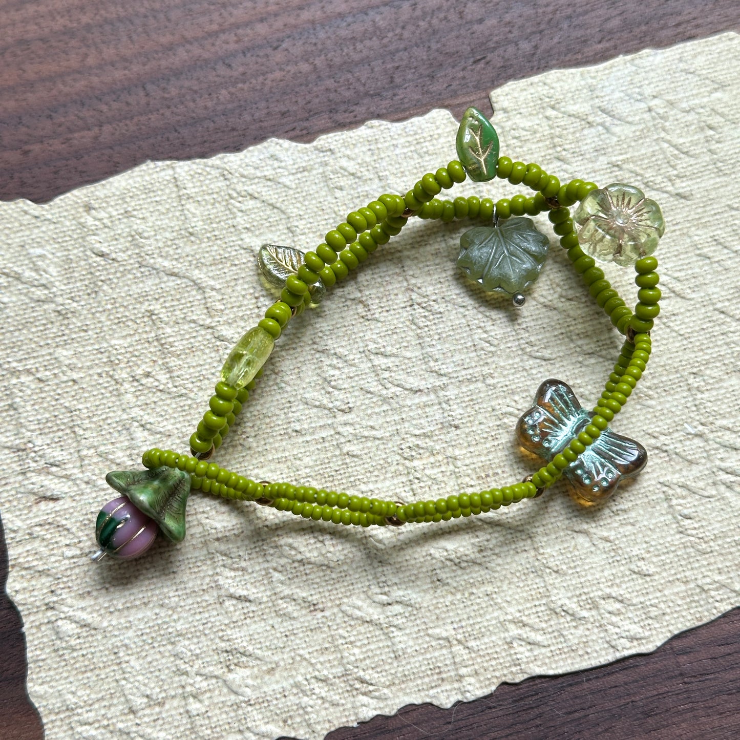 Artisan bracelet with whimsical woodland theme, featuring green beads, delicate leaf details, and a butterfly charm, arranged in circular display.