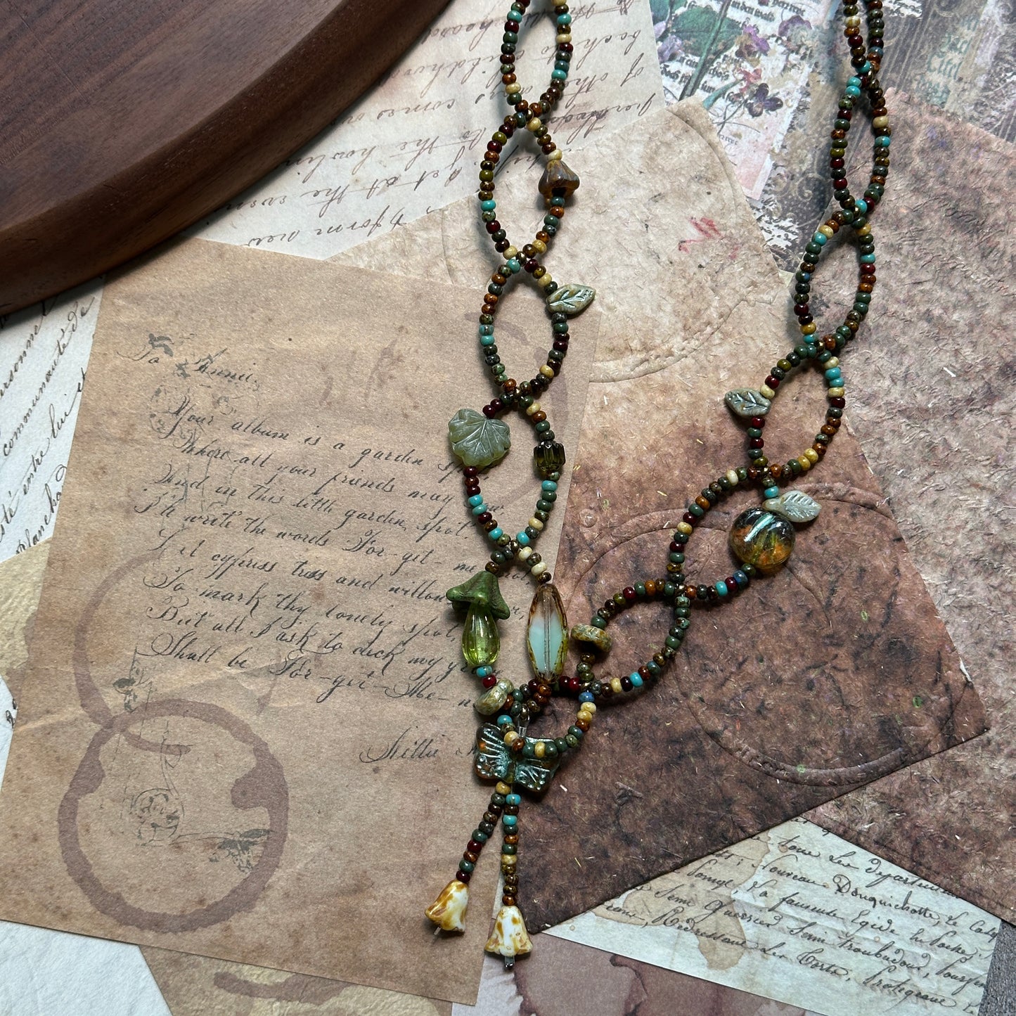 Angled flat lay of the Olive Grove Lariat Necklace showing full length, tassel accents and the woodland olive-amber palette.