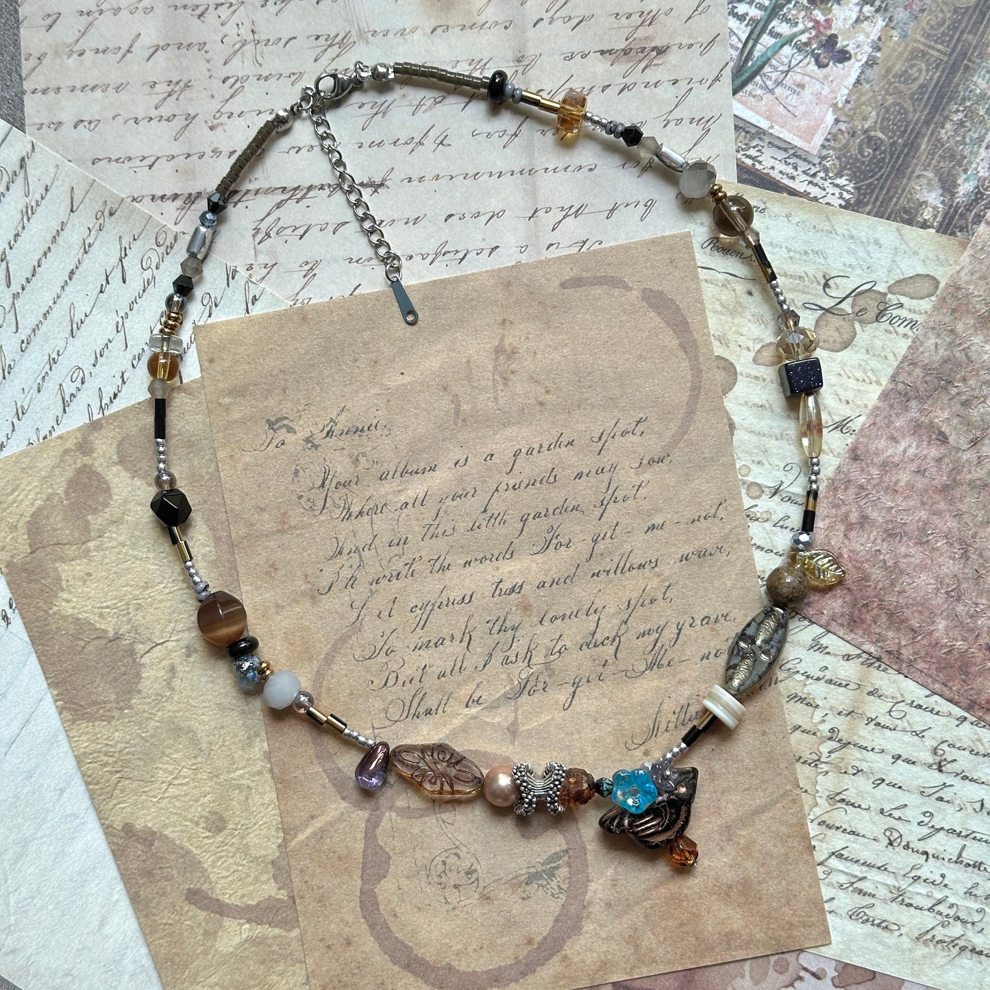 Nomad Relic Necklace coiled flat lay showing matte and faceted beads, slim metal tubes, earth-tone sand/charcoal/bronze palette.