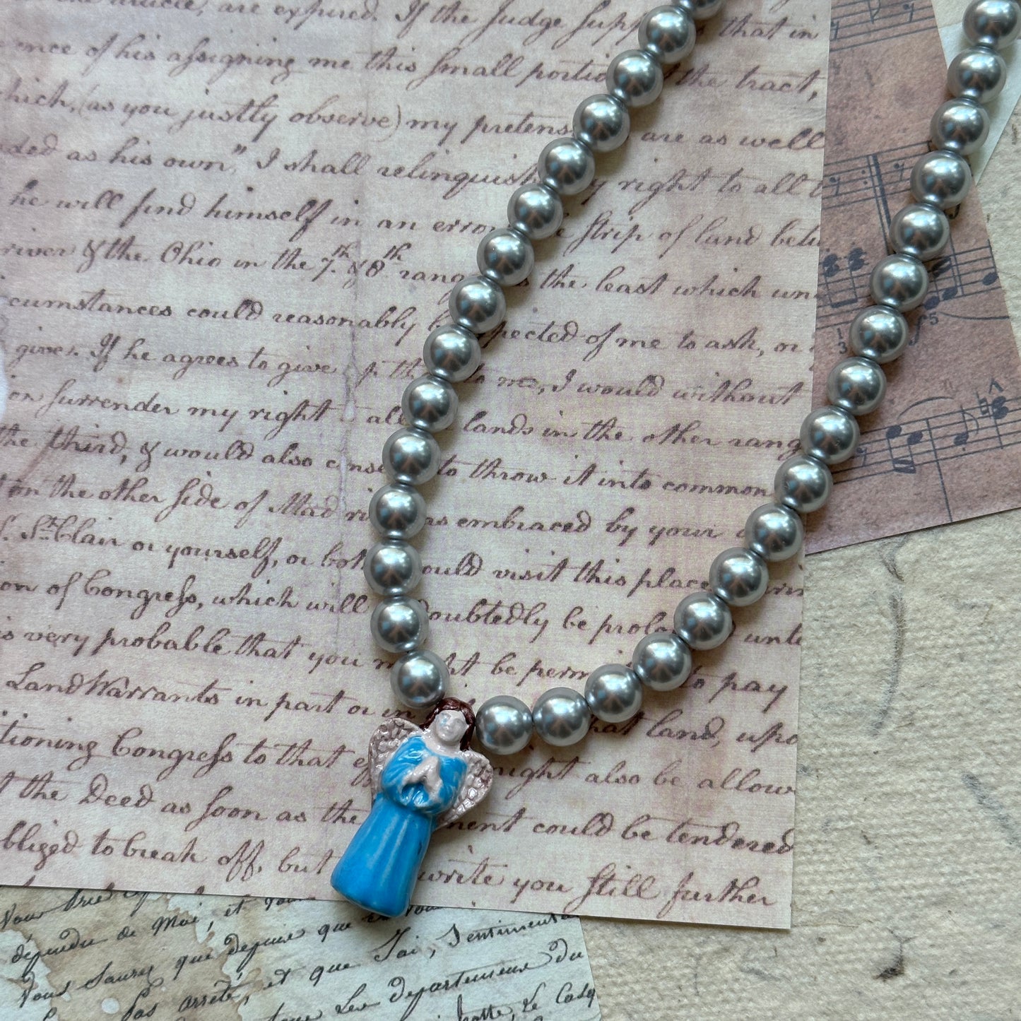 Pendant close-up: sky-blue glass angel charm with silver-sheen faux pearls; elegant, symbolic design.