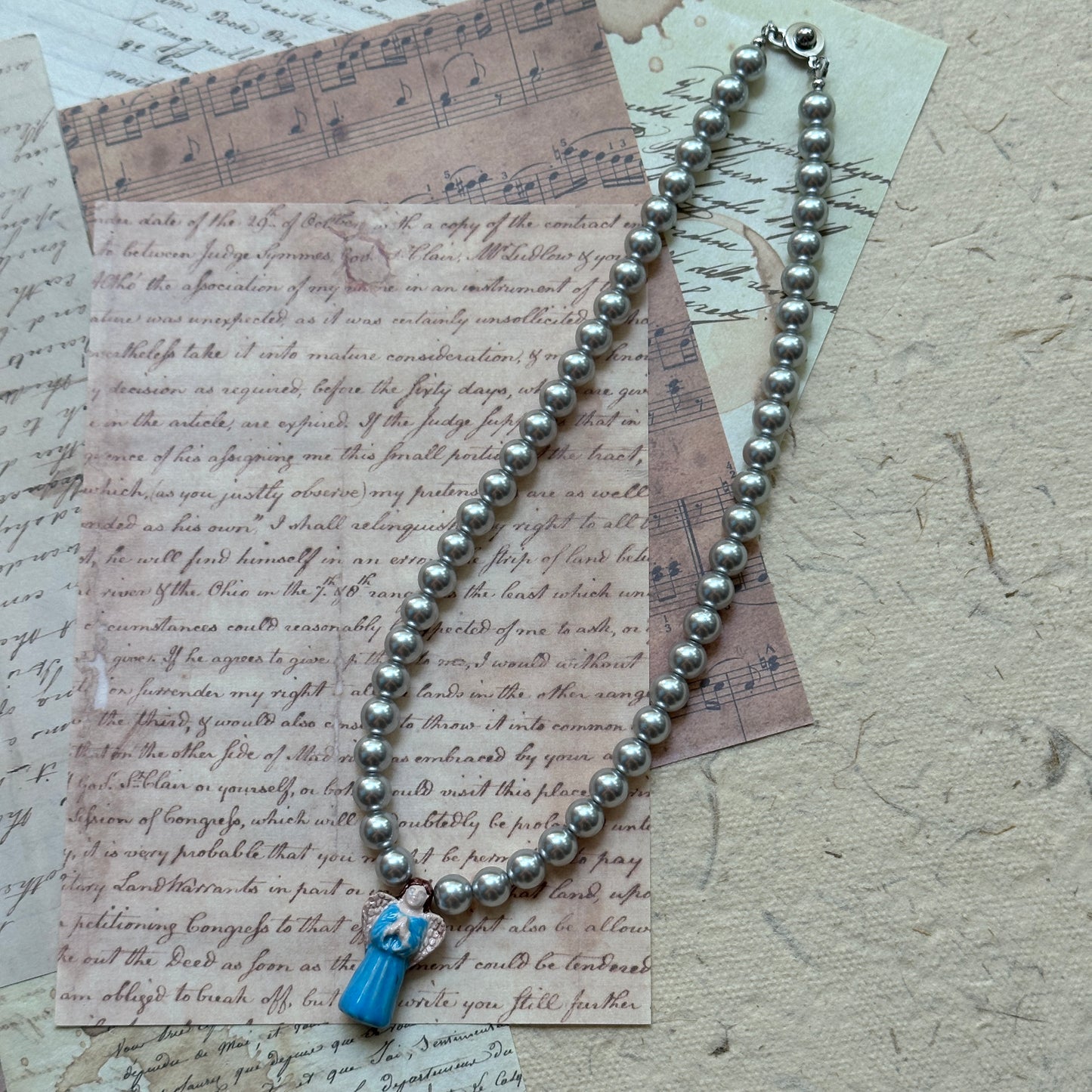 Angled flat lay of the Angel’s Grace Necklace—timeless faux pearls with a delicate blue angel charm.