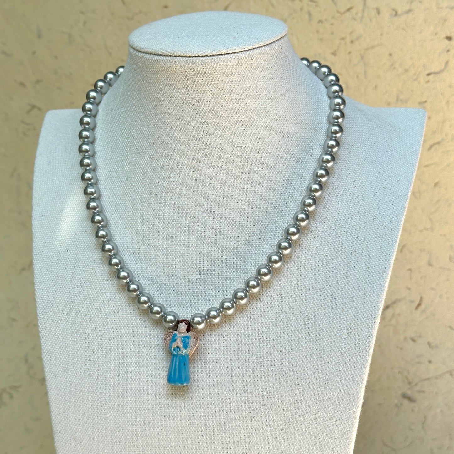 Angel’s Grace Necklace on mannequin—classic faux-pearl strand with sky-blue angel charm; graceful and easy to wear.
