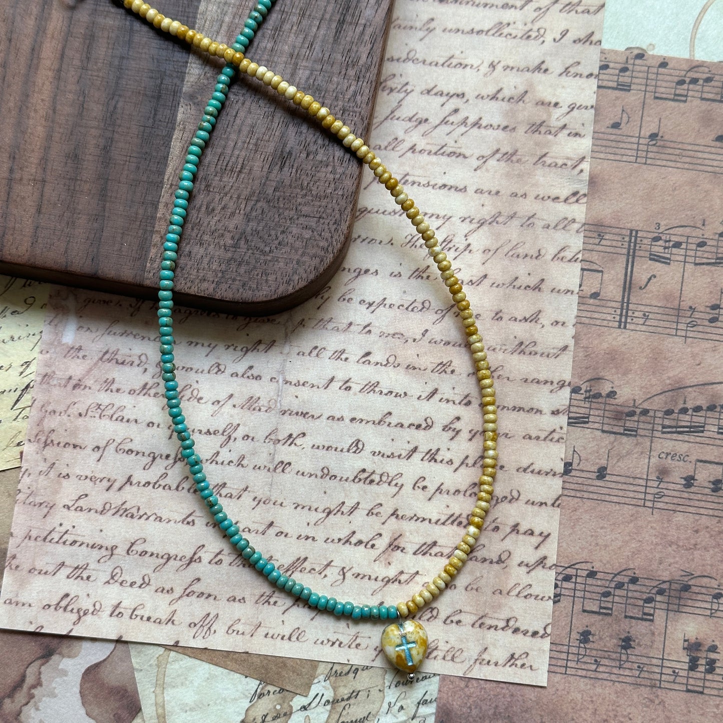 Pendant close-up: small glass cross charm attached to a dual-color strand—amber yellow and teal green; minimal, symbolic design.