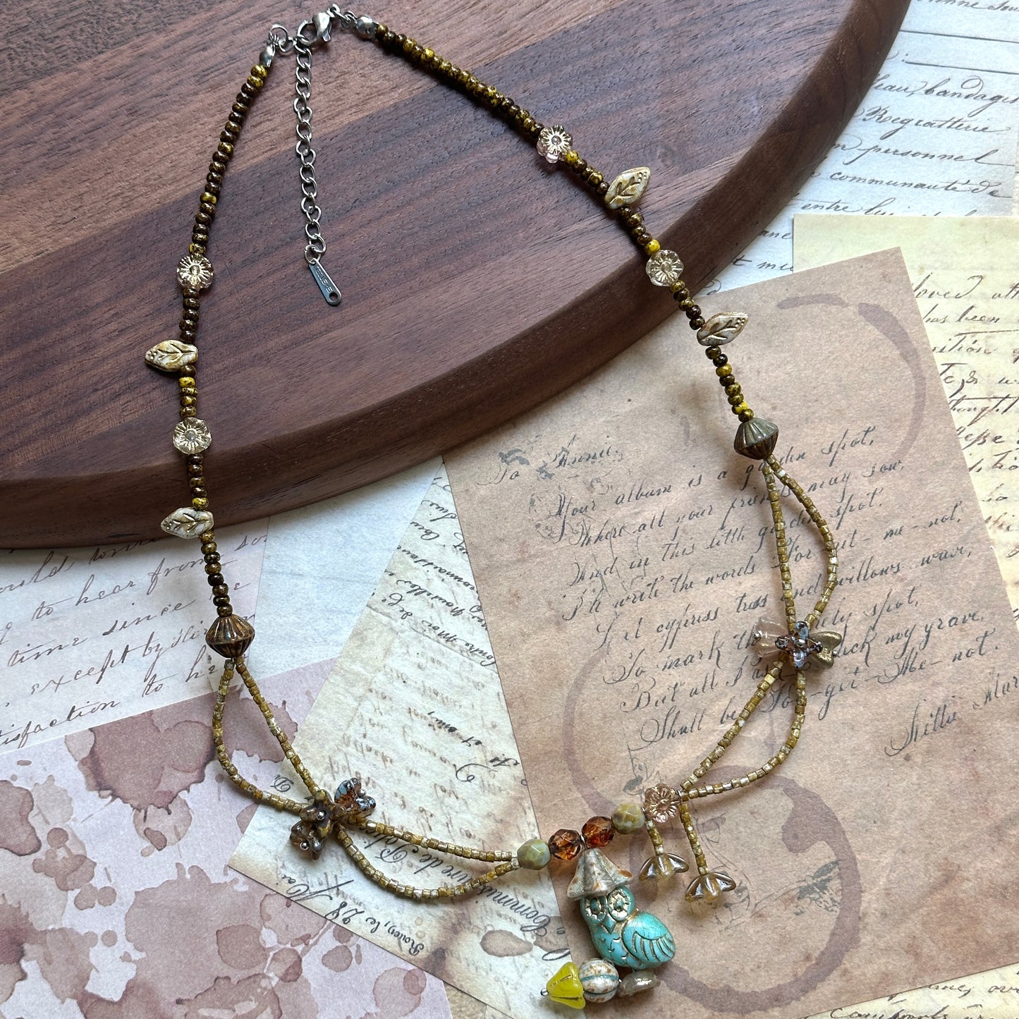 Angled flat lay on parchment—botanical necklace with Czech-glass flower charms and amber beads surrounding a turquoise leaf focal.
