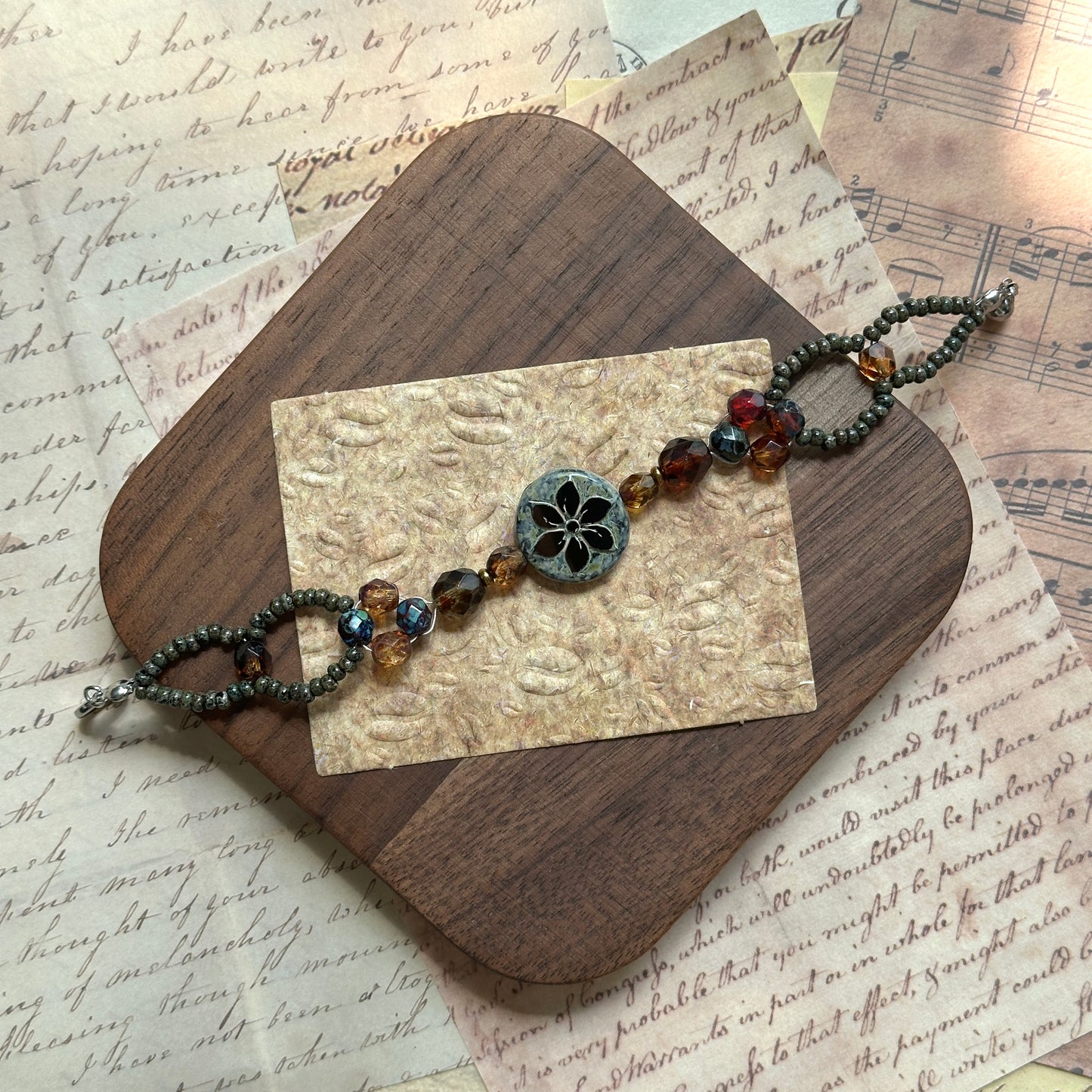 Artisan bracelet styled on a wooden board, floral medallion centerpiece with amber, garnet, and bronze glass beads, elegant oval seed-bead loops