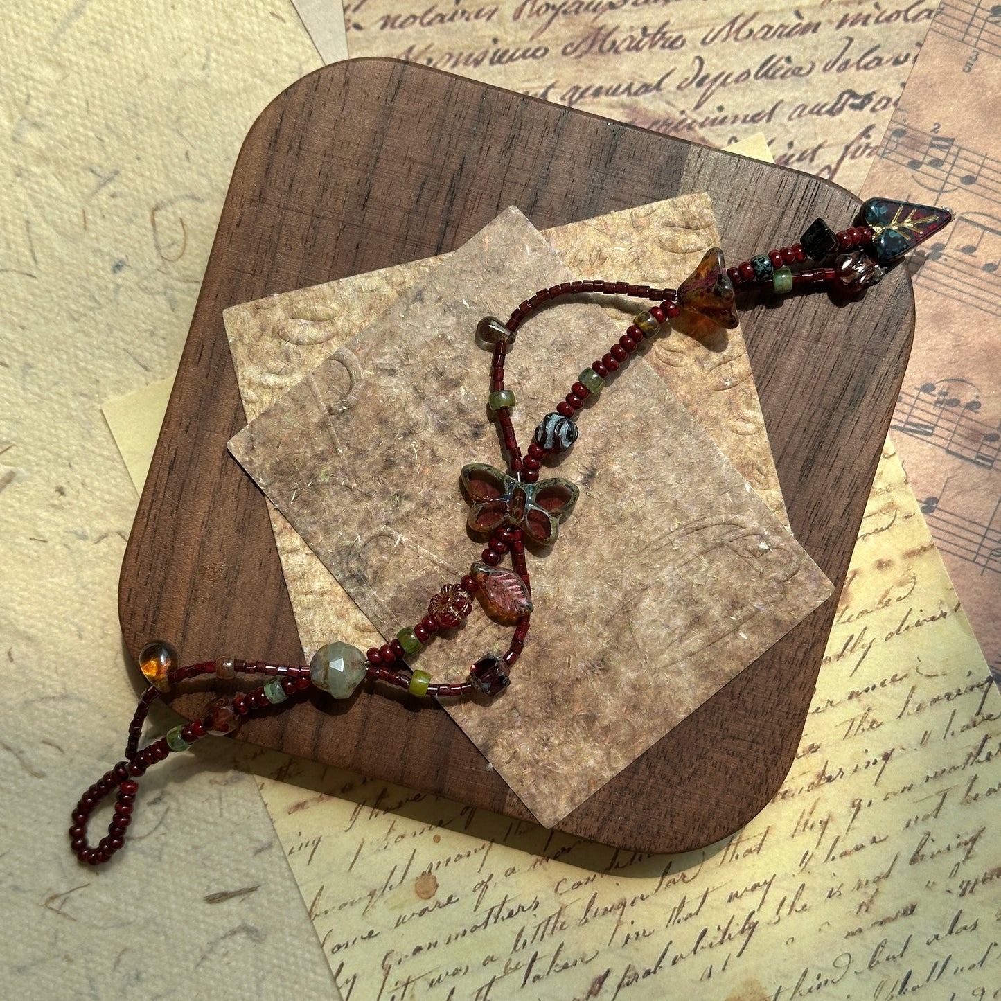 Artisan bracelet styled on a wooden board, deep jewel-tone beads with golden shimmer and leaf details around butterfly focal