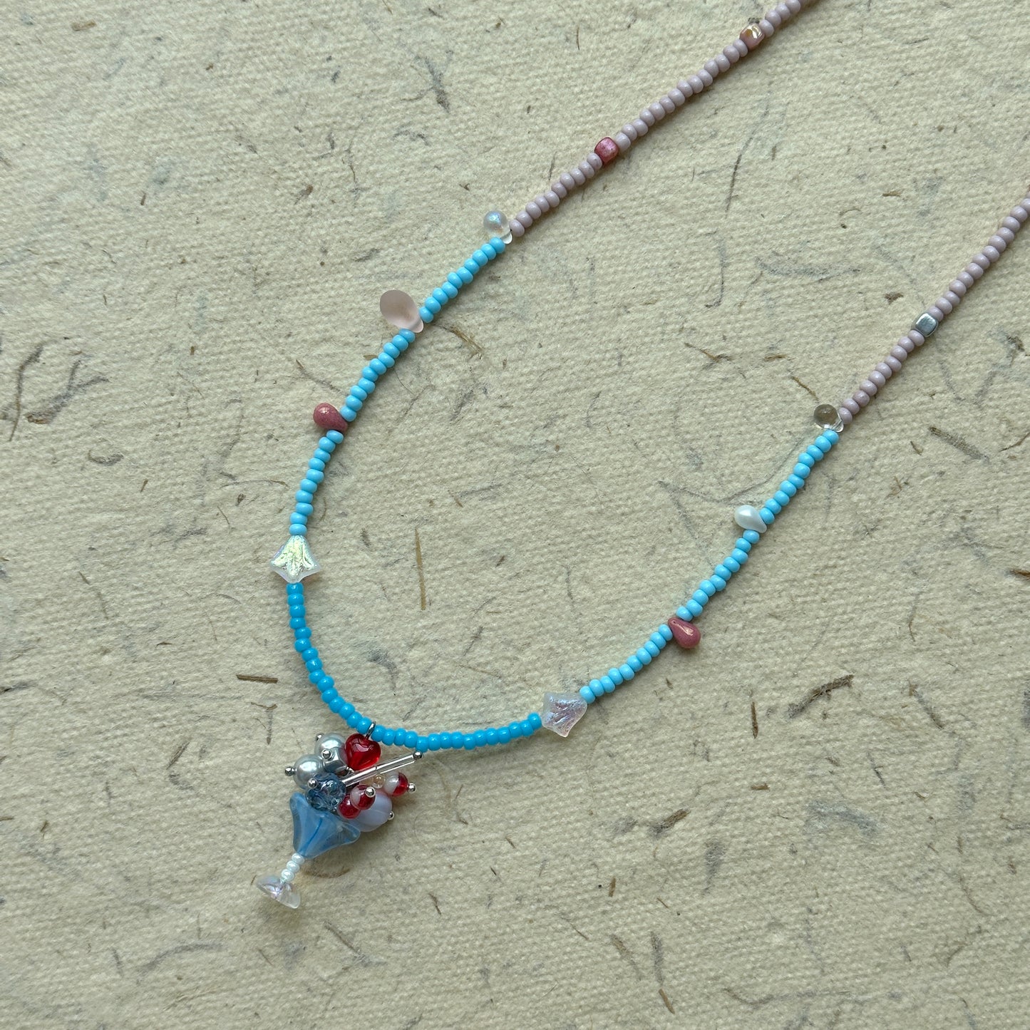 Sky Blossom Charm Necklace flat-lay on fabric, two-tone seed beads in powder pink and sky blue with floral cluster pendant