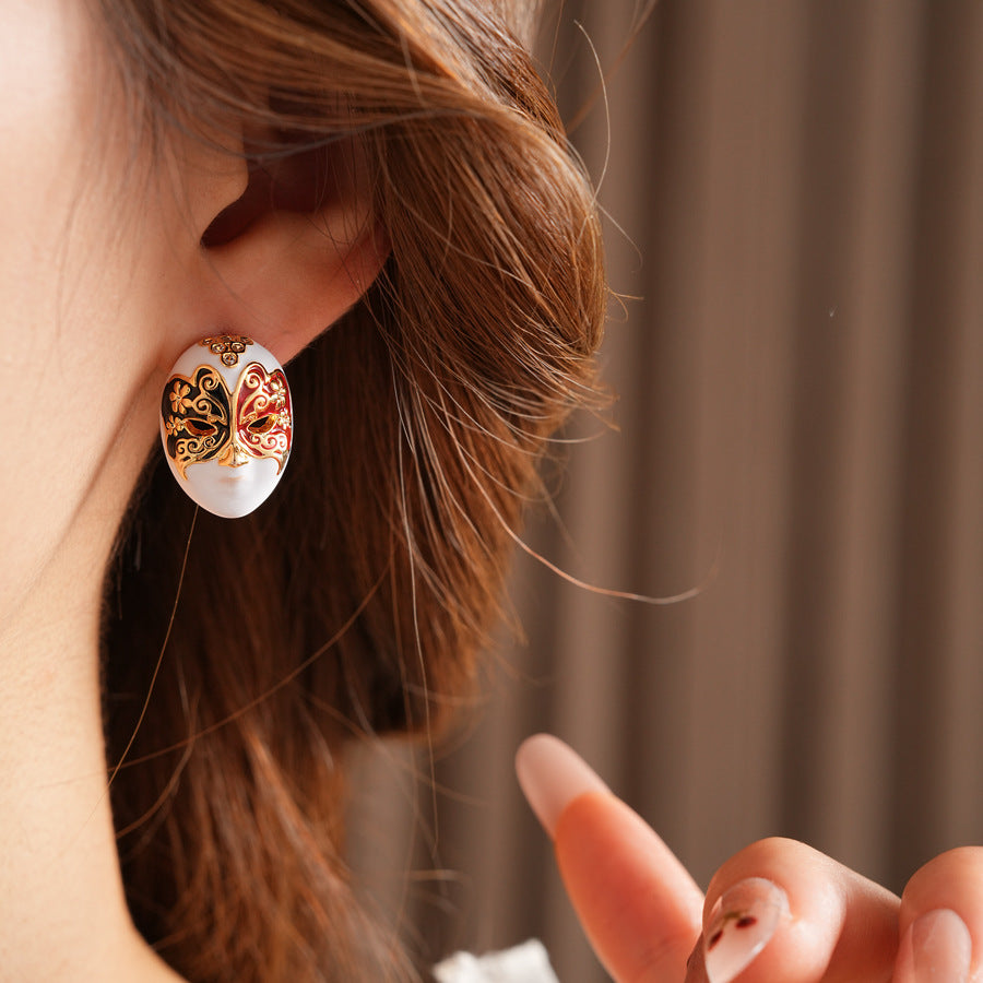 Enamel masquerade face stud earrings with red and black halves and gold filigree on a velvet jewelry box — Venetian Masquerade Studs.