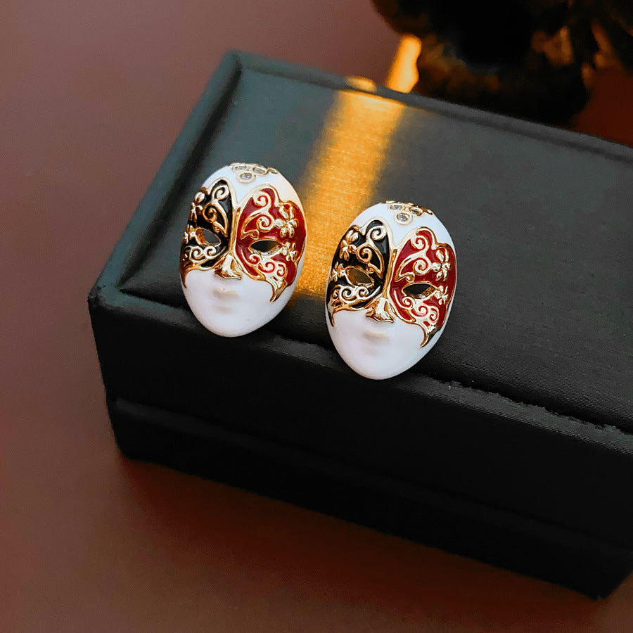Enamel masquerade face stud earrings with red and black halves and gold filigree on a velvet jewelry box — Venetian Masquerade Studs.