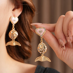 Gold-tone mermaid tail drop earrings with pink enamel heart studs and rhinestone accents on a green velvet background