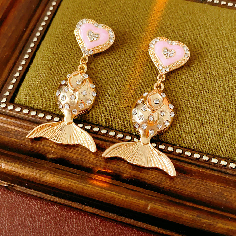 Gold-tone mermaid tail drop earrings with pink enamel heart studs and rhinestone accents on a green velvet background