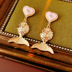 Gold-tone mermaid tail drop earrings with pink enamel heart studs and rhinestone accents on a green velvet background
