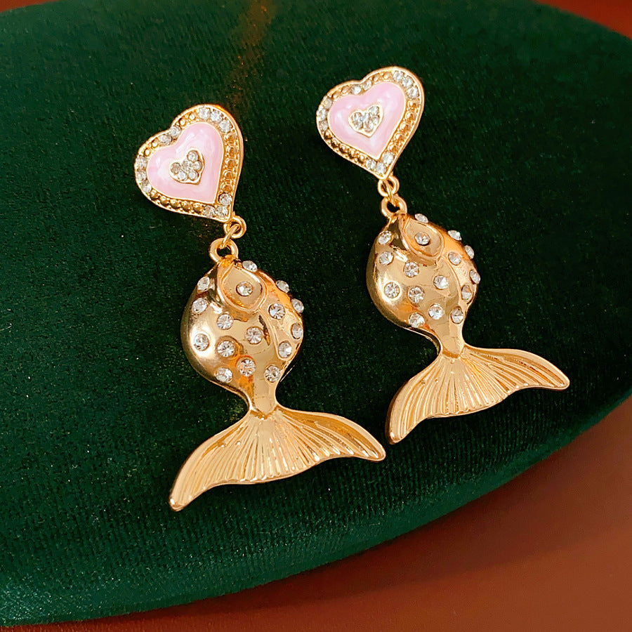 Gold-tone mermaid tail drop earrings with pink enamel heart studs and rhinestone accents on a green velvet background