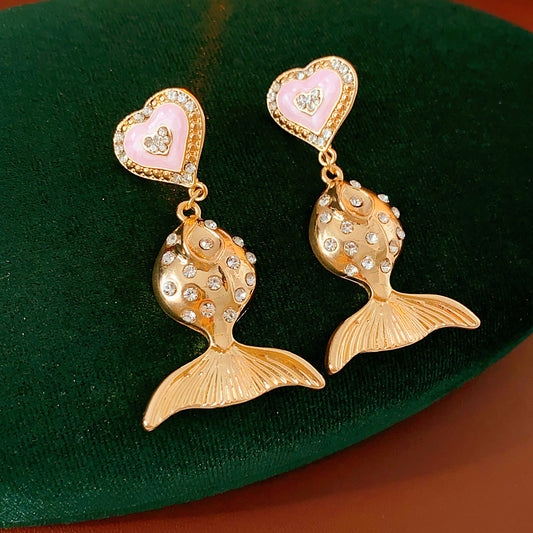 Gold-tone mermaid tail drop earrings with pink enamel heart studs and rhinestone accents on a green velvet background