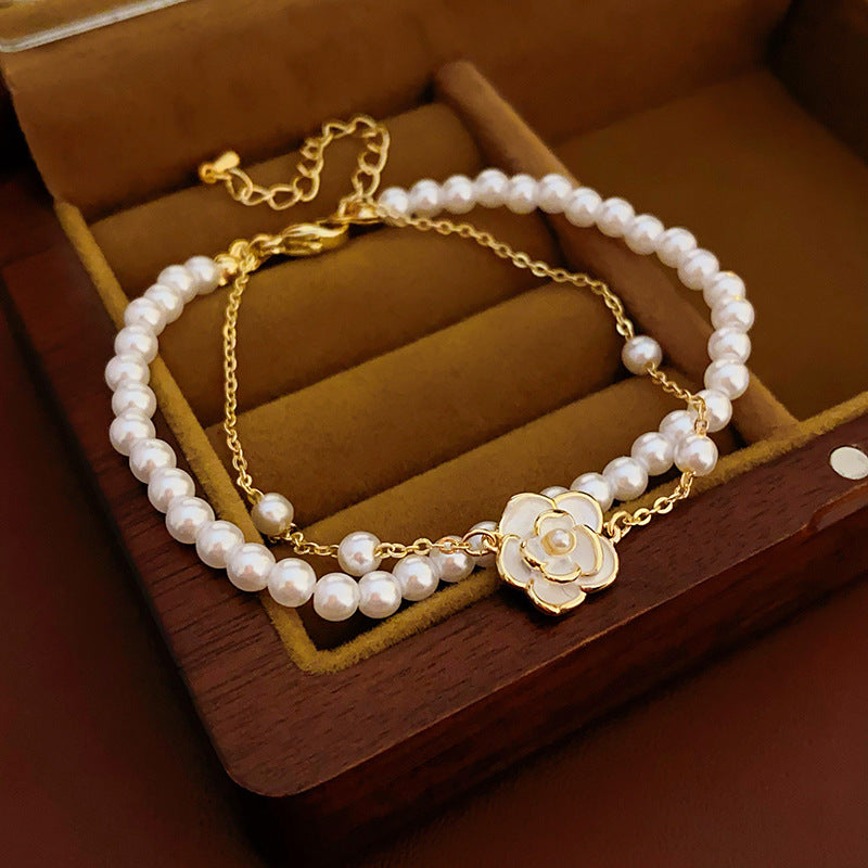 Layered faux-pearl bracelet with gold-tone chain and white enamel flower charm placed in a green jewelry tray — Petite Pearl Bloom Layered Bracelet.
