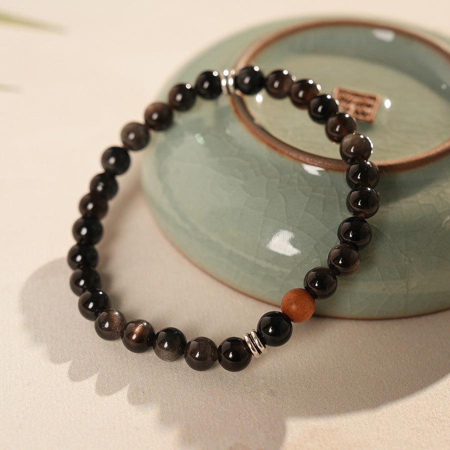 Sleek black silver-sheen stone beaded bracelet with one warm wood bead and silver ring spacers on a ceramic dish — Shadowwood Sheen Bracelet.