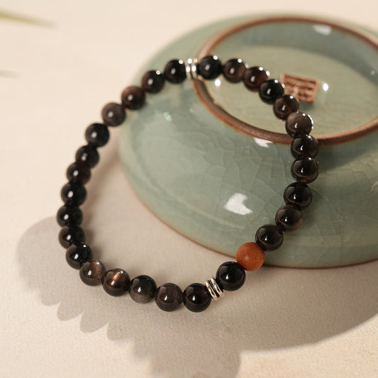 Sleek black silver-sheen stone beaded bracelet with one warm wood bead and silver ring spacers on a ceramic dish — Shadowwood Sheen Bracelet.