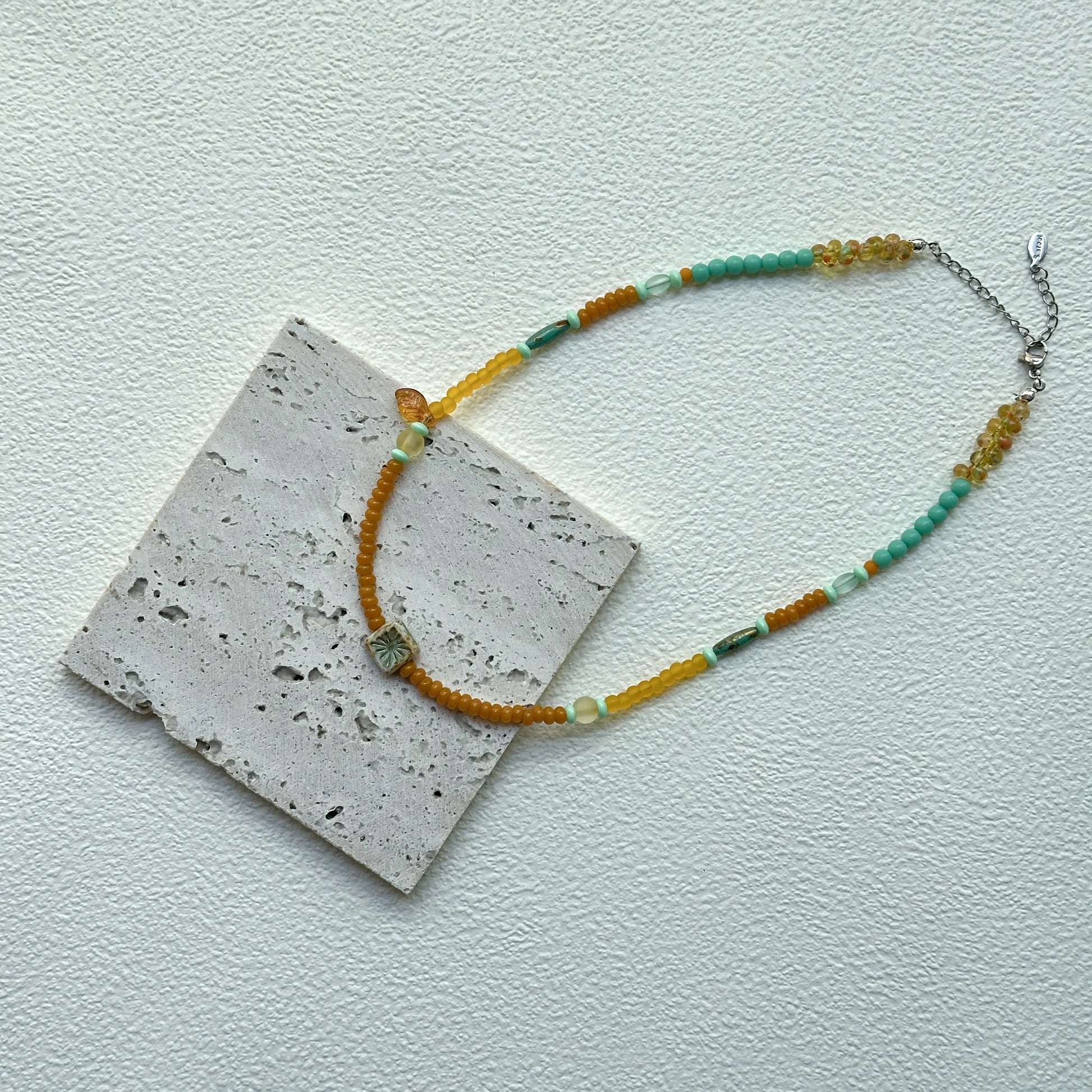 Flat lay of Citrus Lagoon Necklace on textured card, full view with floral tile centerpiece and small amber leaf accent