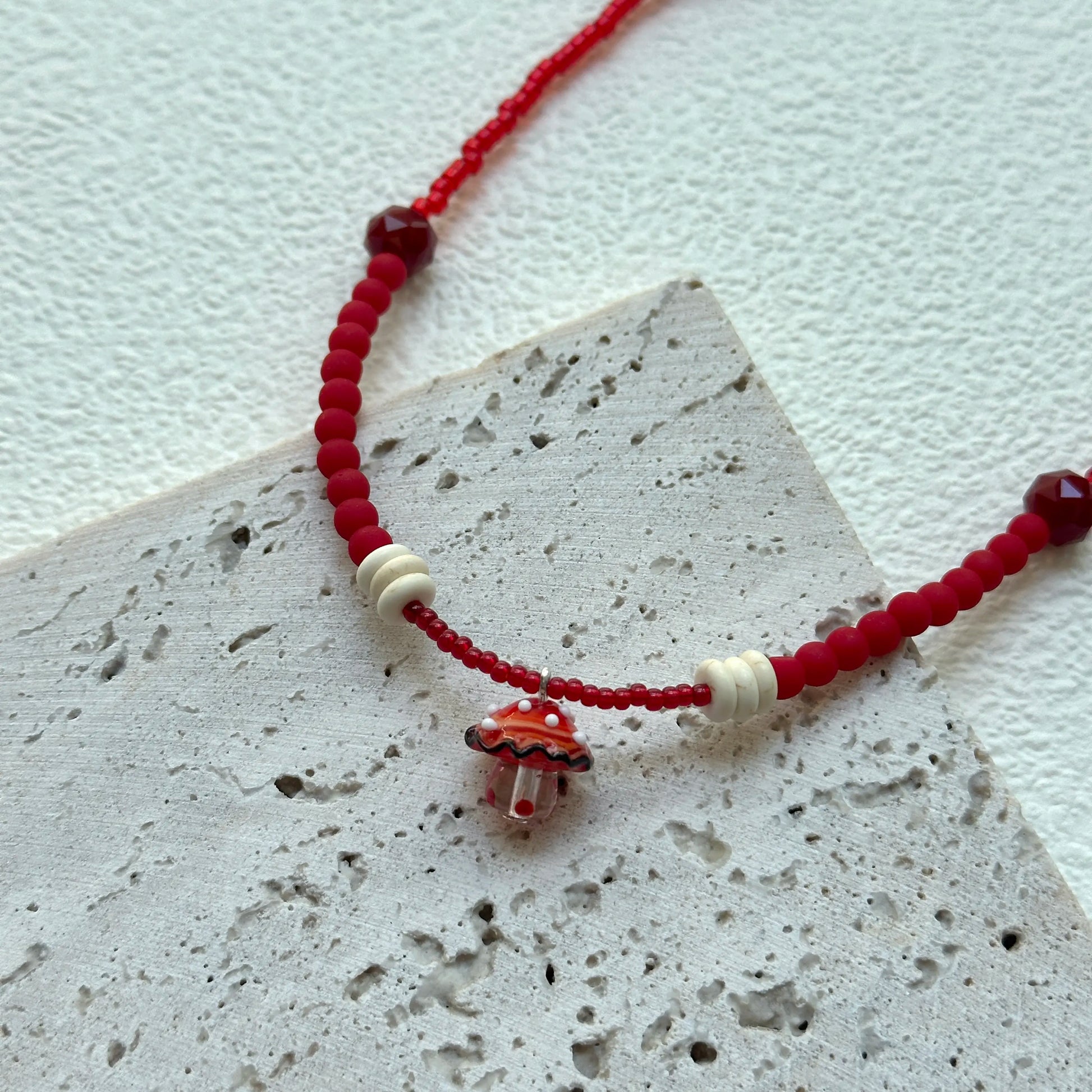 Crimson Toadstool Necklace styled on textured card, close-up of lampwork mushroom pendant with red and ivory beads