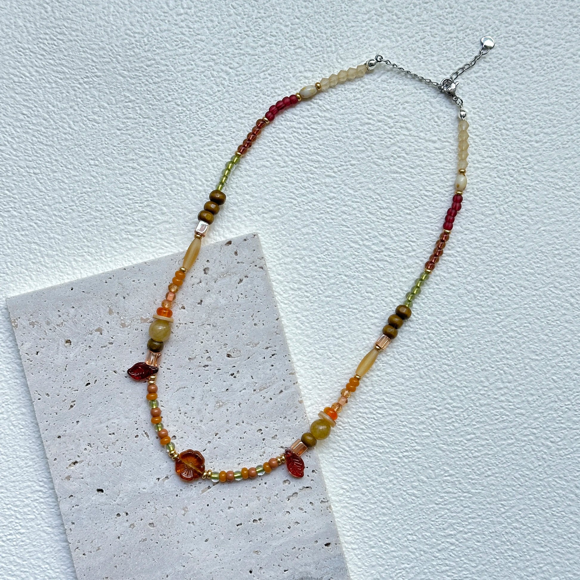 Flat lay of Olive Grove Blossom Necklace on textured card – handcrafted olive/amber palette with floral centerpiece and petite leaf details