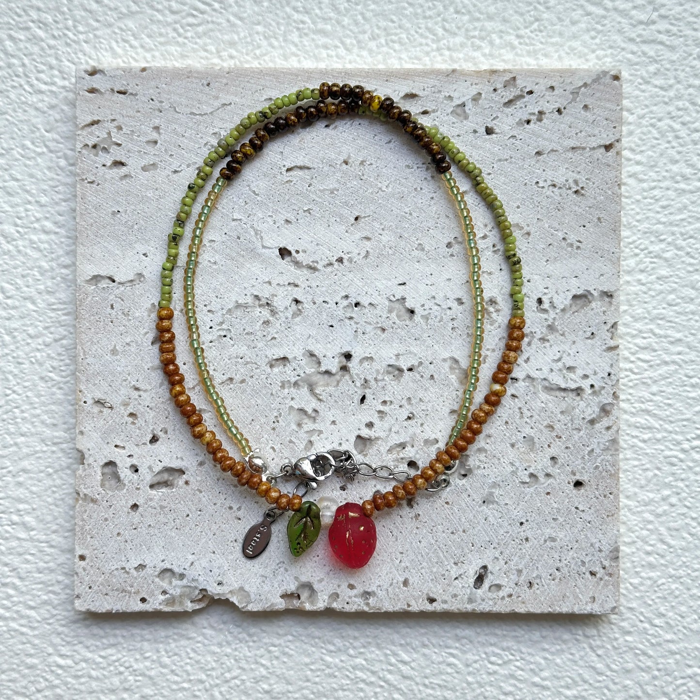Wild Strawberry Necklace styled on a textured card, close-up of strawberry focal with leaf and dewdrop accents – handmade by Glazenya