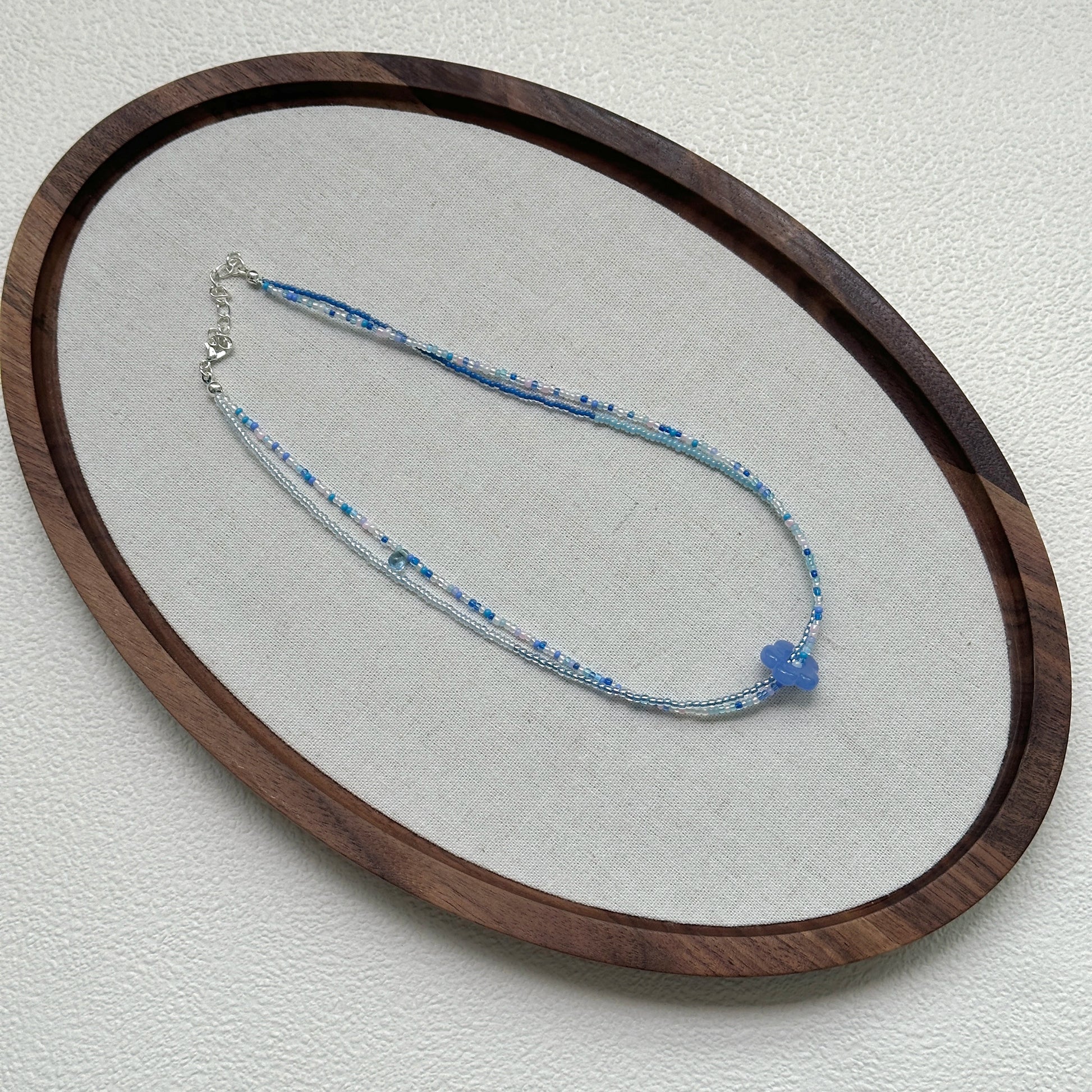 Azure Bloom Necklace oval flat lay – handcrafted minimal beaded necklace with azure flower charm and mixed blue-white beads by Glazenya