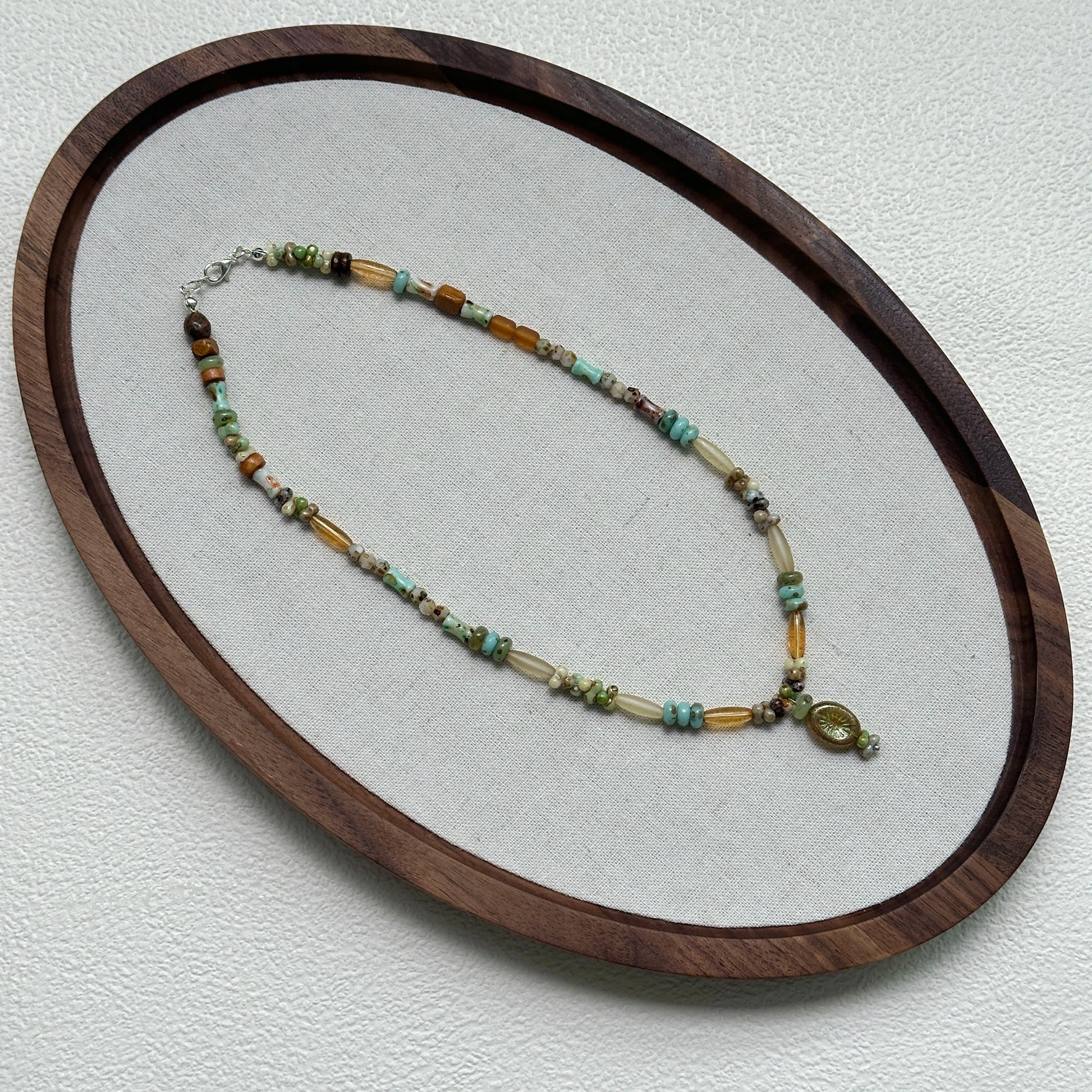 Verdant Amulet Necklace oval flat lay – handcrafted beaded necklace with amber-gold, green, and ivory beads surrounding a vintage pendant by Glazenya