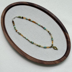 Verdant Amulet Necklace oval flat lay – handcrafted beaded necklace with amber-gold, green, and ivory beads surrounding a vintage pendant by Glazenya