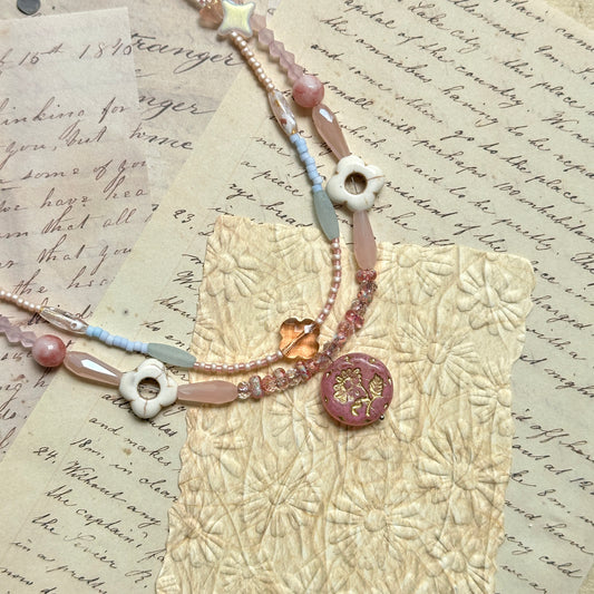 Sakura Medallion Necklace — close-up on vintage papers; double-strand pastel beads in blush, pearl-white and sky-blue with heart accents and a sakura medallion charm