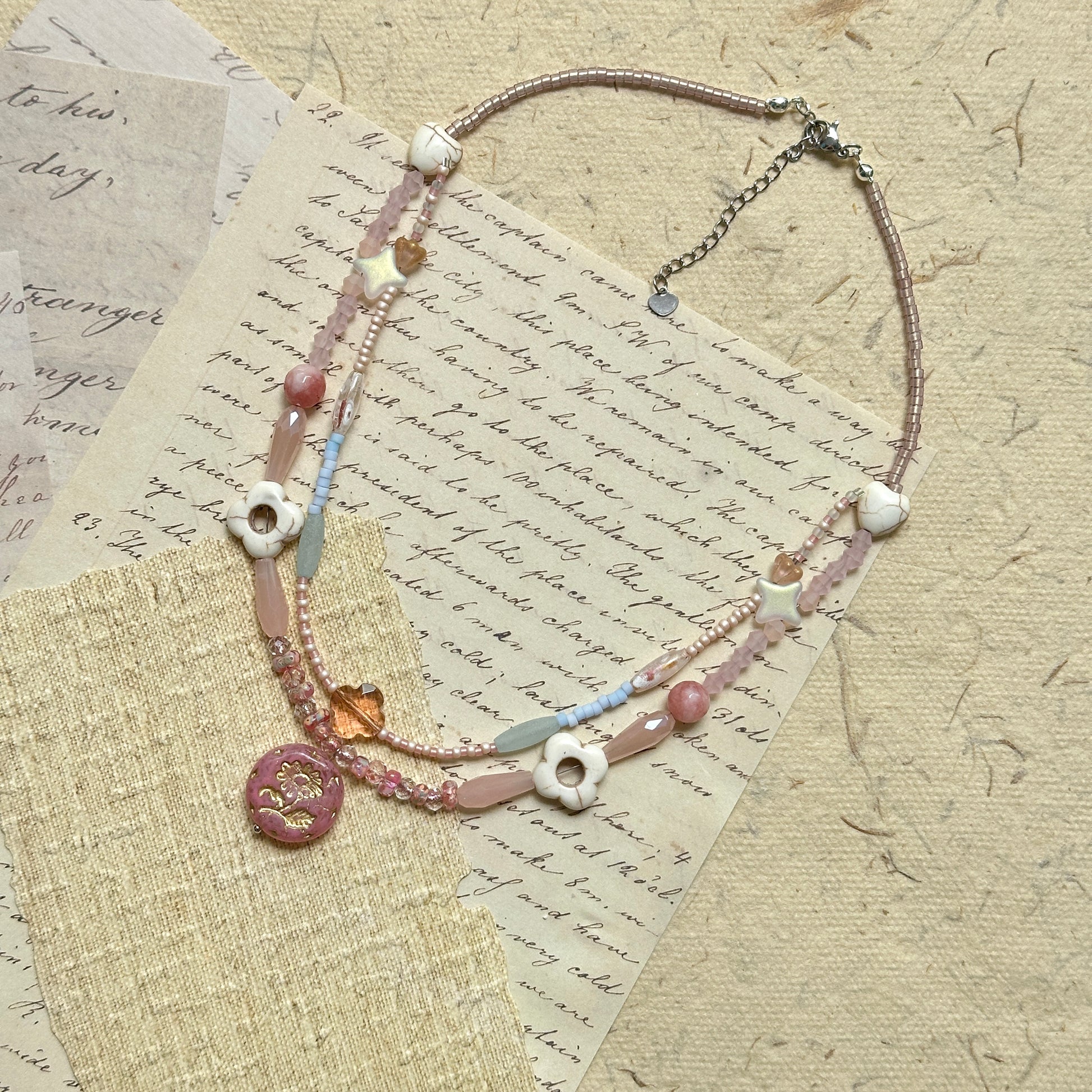 Sakura Medallion Necklace — flat lay top-down on textured paper; full double-strand with heart/petal accents and sakura medallion focal