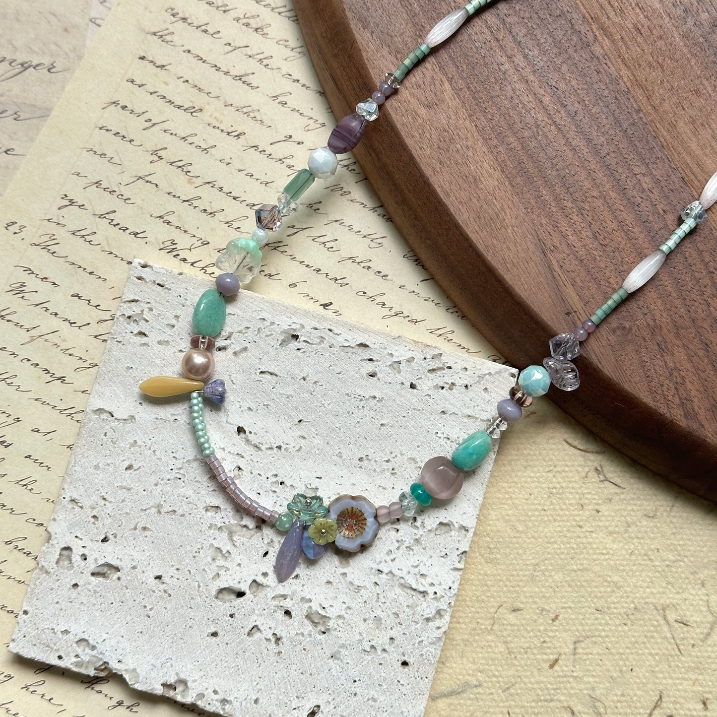 Seafoam Petal Necklace — close-up flat lay on textured papers beside a round wooden tray; pastel seafoam, lilac and pearl-white beads with floral and petal-shaped accents
