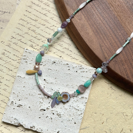 Seafoam Petal Necklace — close-up flat lay on textured papers beside a round wooden tray; pastel seafoam, lilac and pearl-white beads with floral and petal-shaped accents