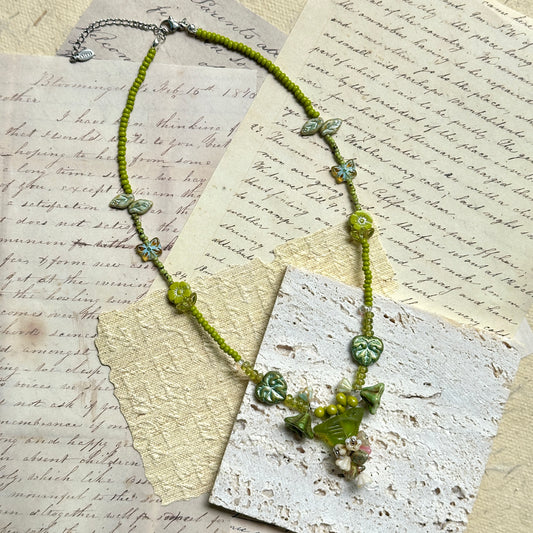 Limeleaf Bouquet Necklace — flat lay on vintage papers; lime-green seed beads with leaf and butterfly accents and a bouquet-style focal charm