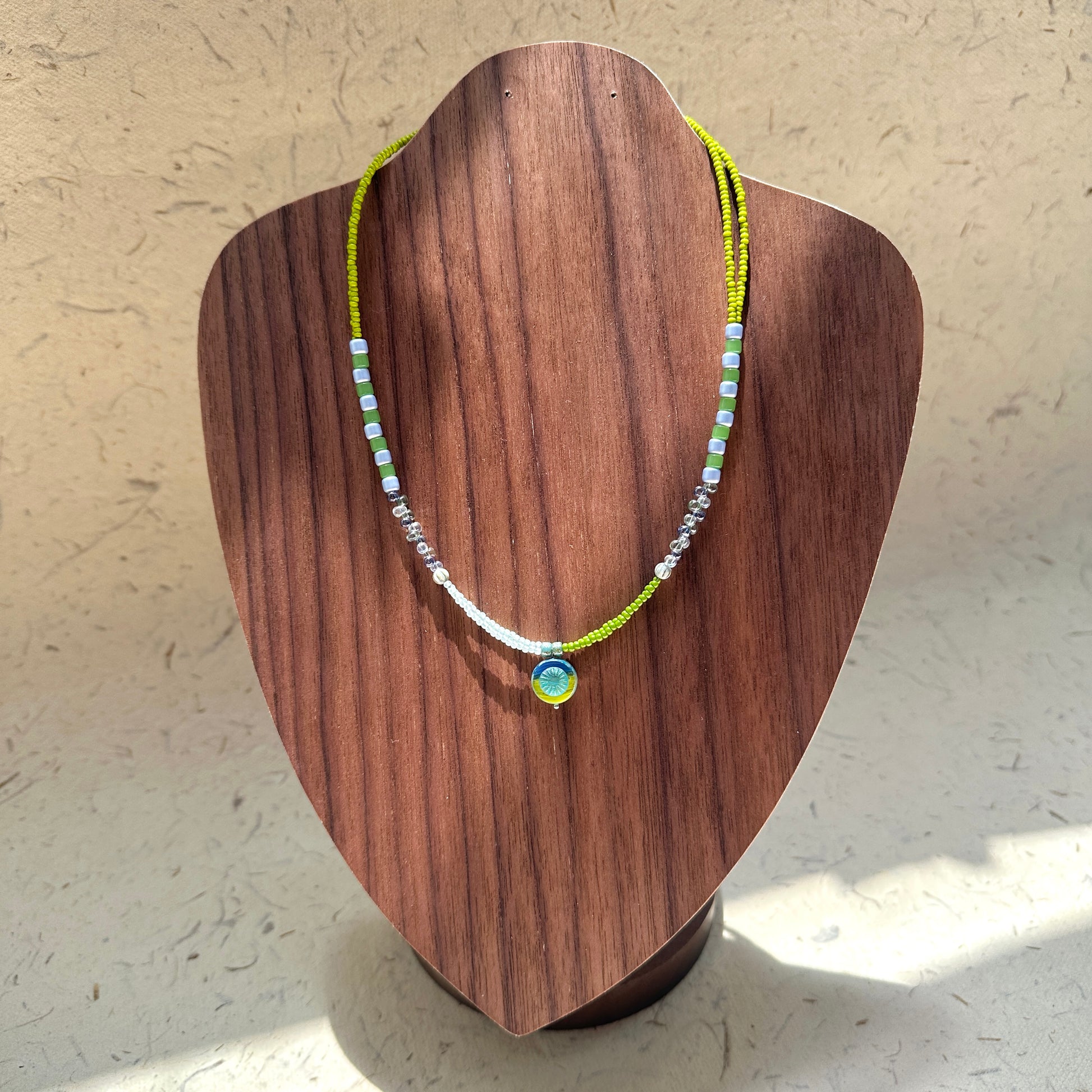 Citrus Wave Necklace on wood display bust — lime and aqua beads with striped segments and a small wave medallion pendant