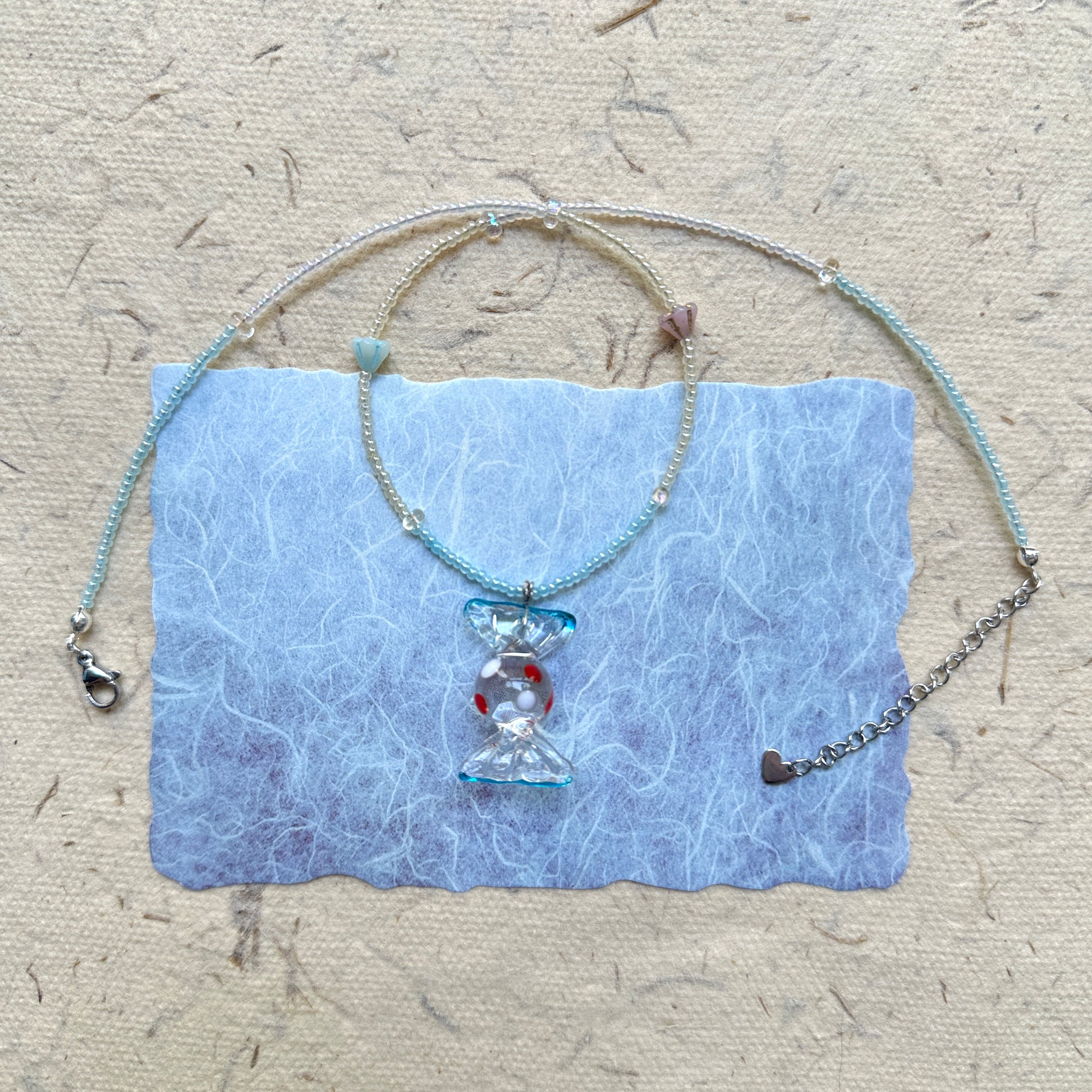Ocean Bonbon Necklace — flat lay on textured paper; aqua and pearl beads with a glass candy pendant featuring red polka dots
