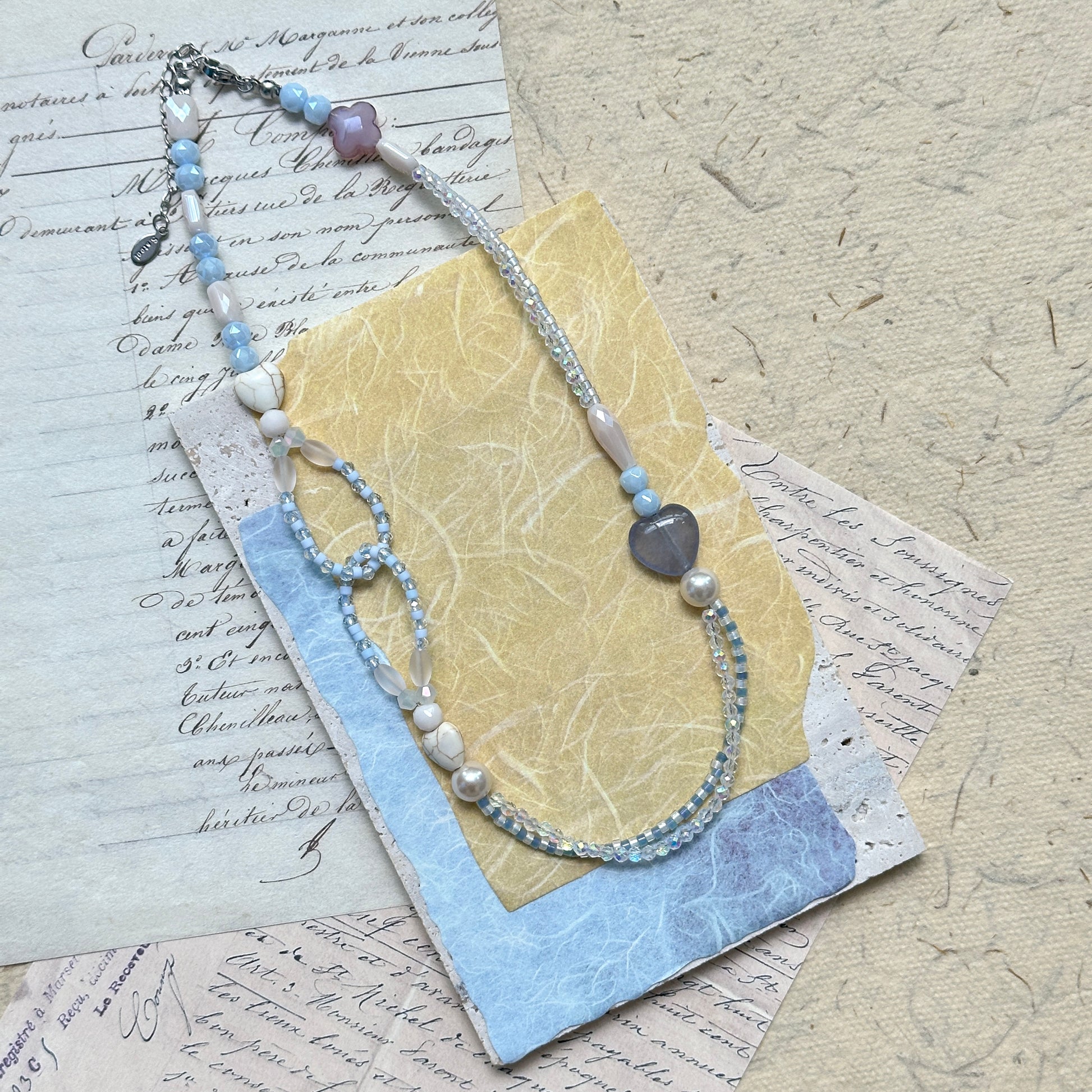 Lunar Mist Necklace — flat lay on textured paper; asymmetric double-strand in icy blue and pearl white with heart and loop details