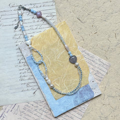 Lunar Mist Necklace — flat lay on textured paper; asymmetric double-strand in icy blue and pearl white with heart and loop details
