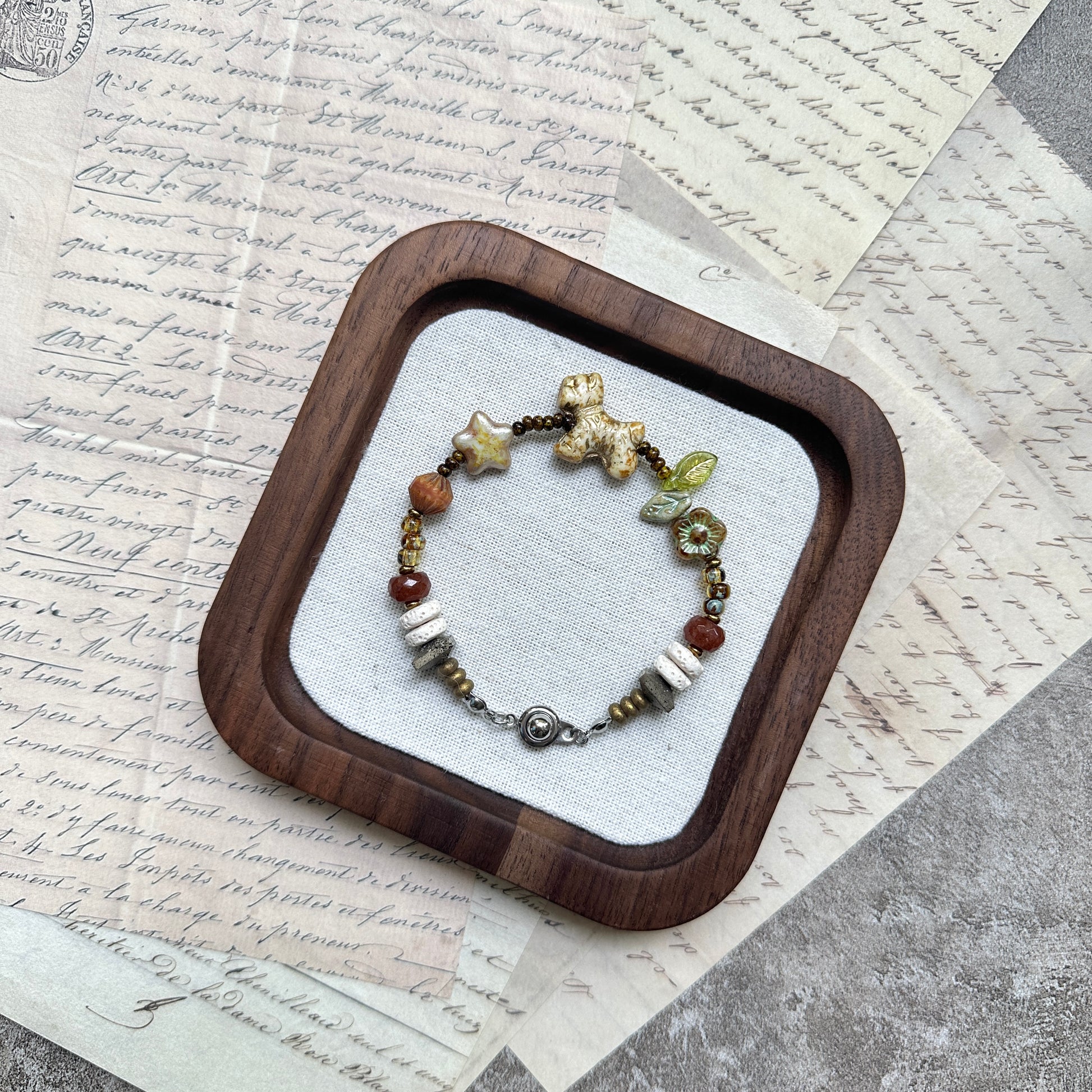 Meadow Pup Bracelet — flat lay in a wooden tray on vintage papers; warm amber and neutral beads with puppy, leaf, star, and floral accents