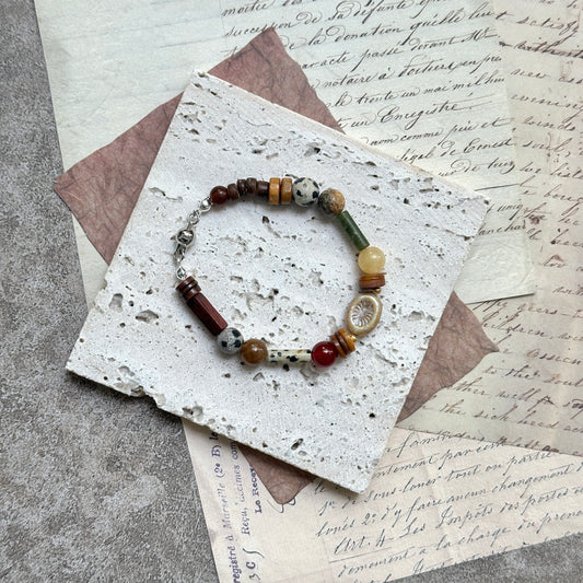 Spice Caravan Bracelet — flat lay in a wooden tray on vintage papers; saffron, amber and olive-green beads with a sunburst oval charm