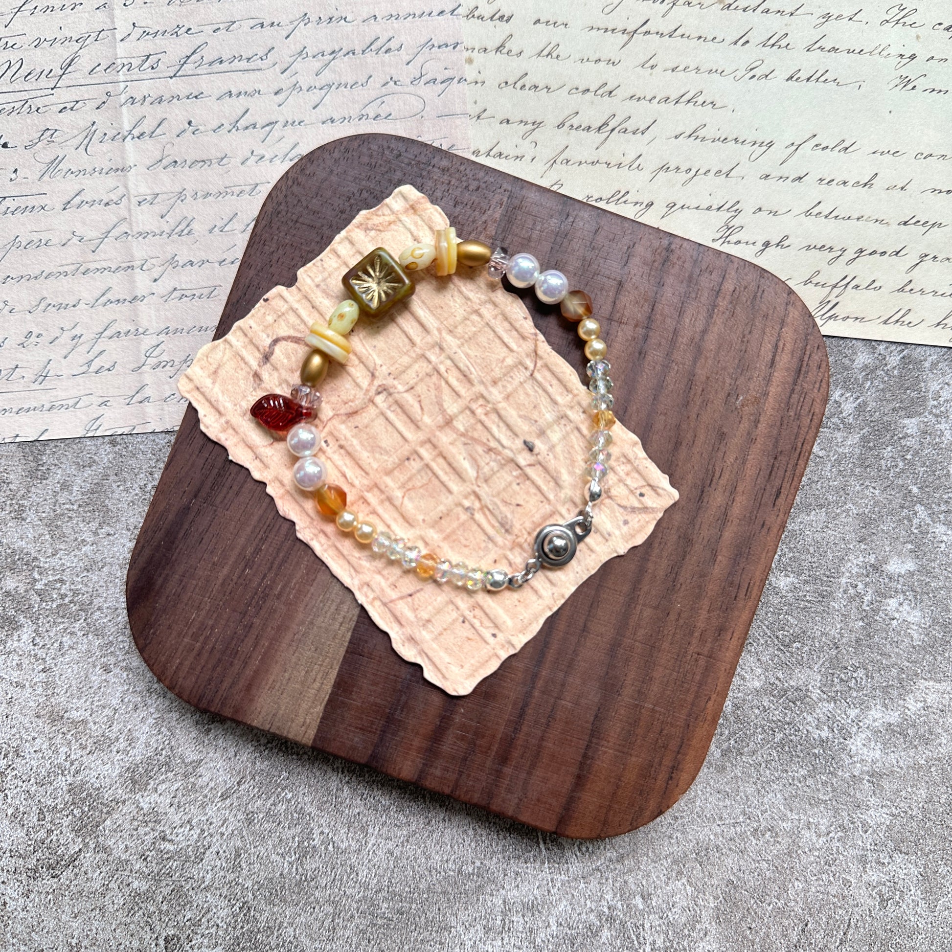 Olive Sunburst Bracelet — flat lay in a wooden tray on textured papers; honey and pearl beads with an olive square sunburst charm, red leaf accent, and magnetic clasp